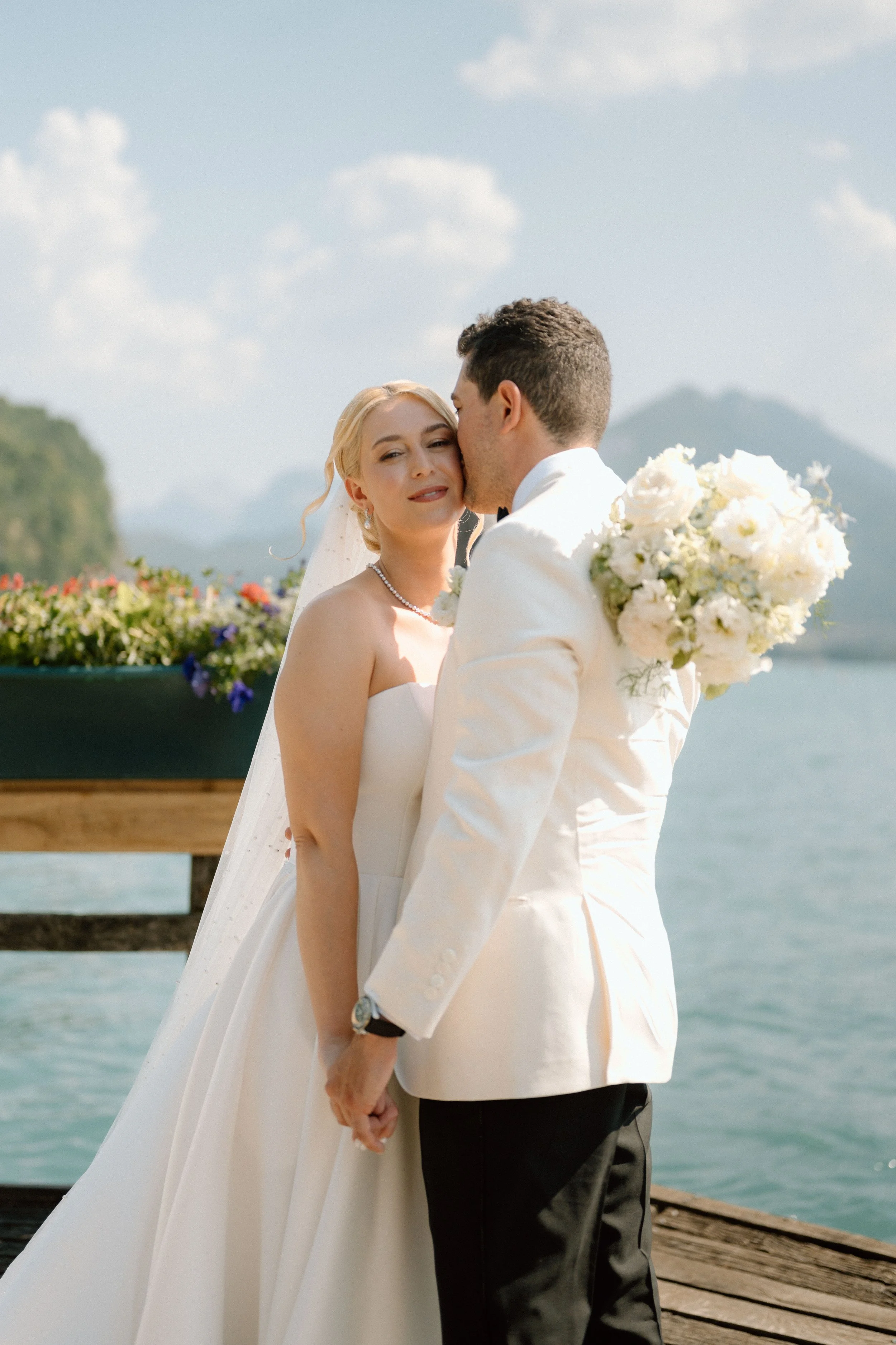 Un couple en mariage échangeant un baiser sur une jetée en bois au bord de l'eau avec un décor de montagne en arrière-plan, lors d'une journée ensoleillée.