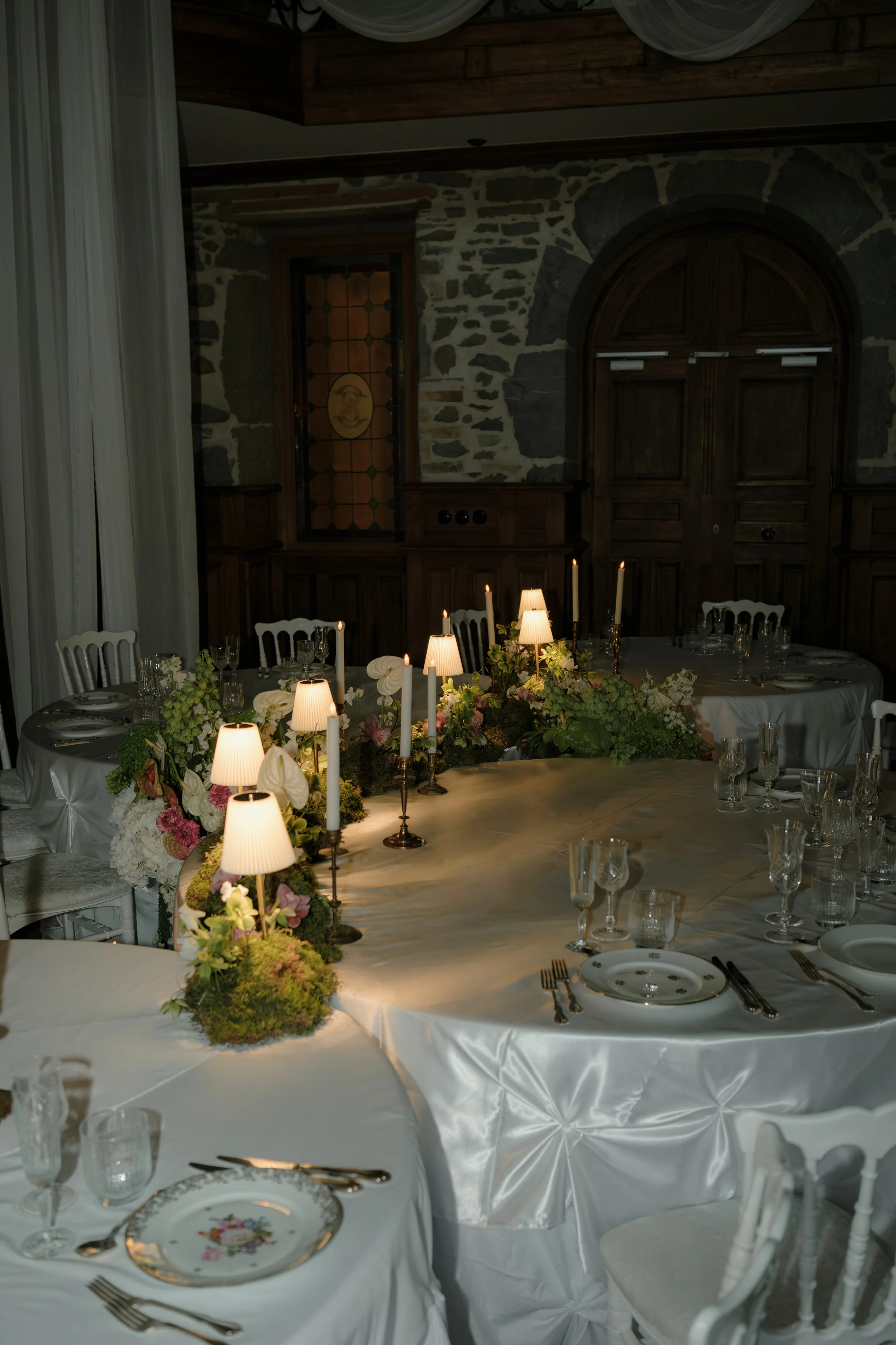Table de réception élégante décorée avec des fleurs, des bougies et de la vaisselle raffinée dans une salle avec murs en pierre et fenêtres en bois.