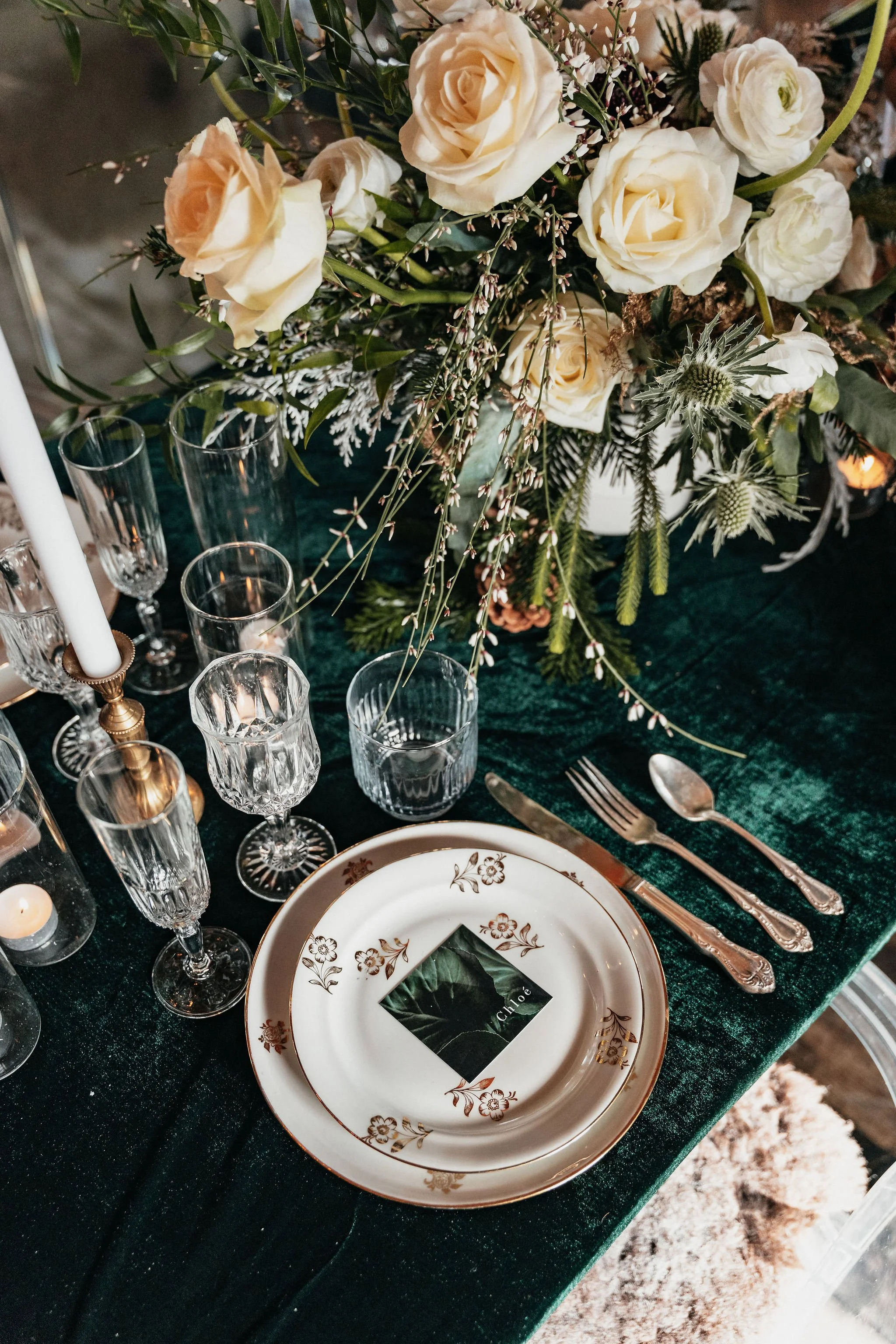 Table élégante décorée avec un grand bouquet de fleurs blanches et crème, des verres à vin en cristal, une assiette en porcelaine blanche avec motifs dorés, une petite carte avec le nom 'Chloé', et des bougies.