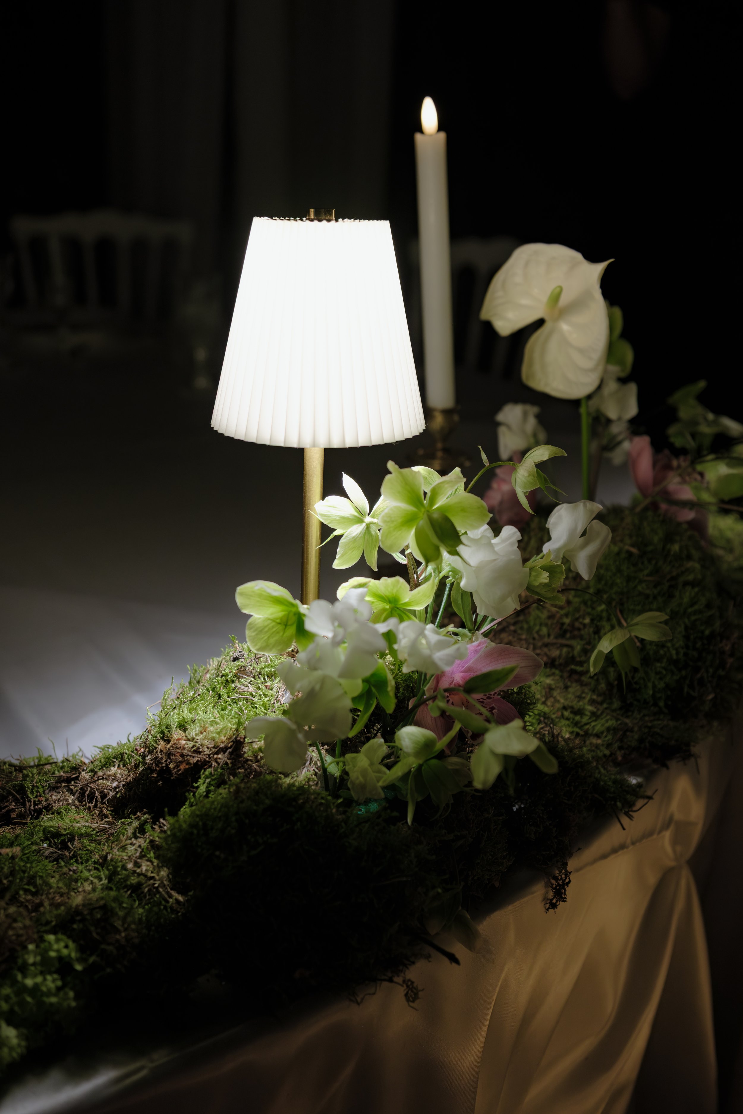 Une table décorée avec une lampe à abat-jour blanc, une bougie candle fine allumée, et un arrangement floral de différentes fleurs blanches et vertes sur un fond sombre.
