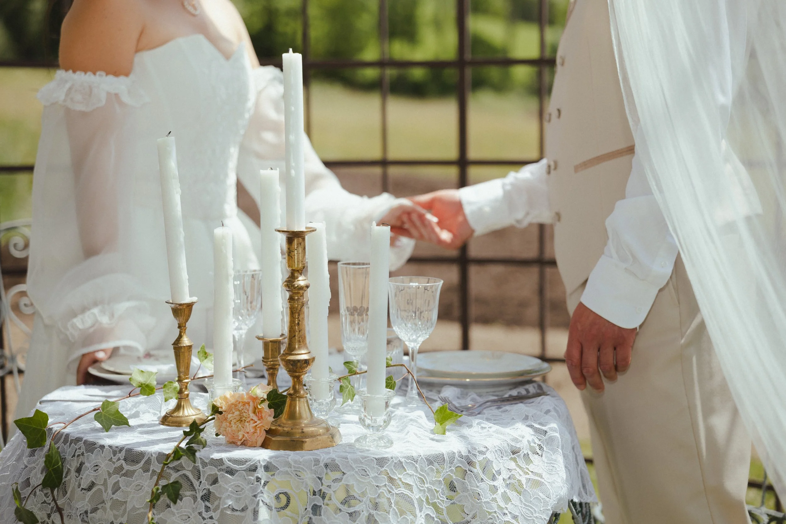 Un couple lors d'une cérémonie nuptiale échangeant une poignée de main, avec une table décorée de bougies blanches, de fleurs pastel et de feuillage vert, sur une terrasse en extérieur.