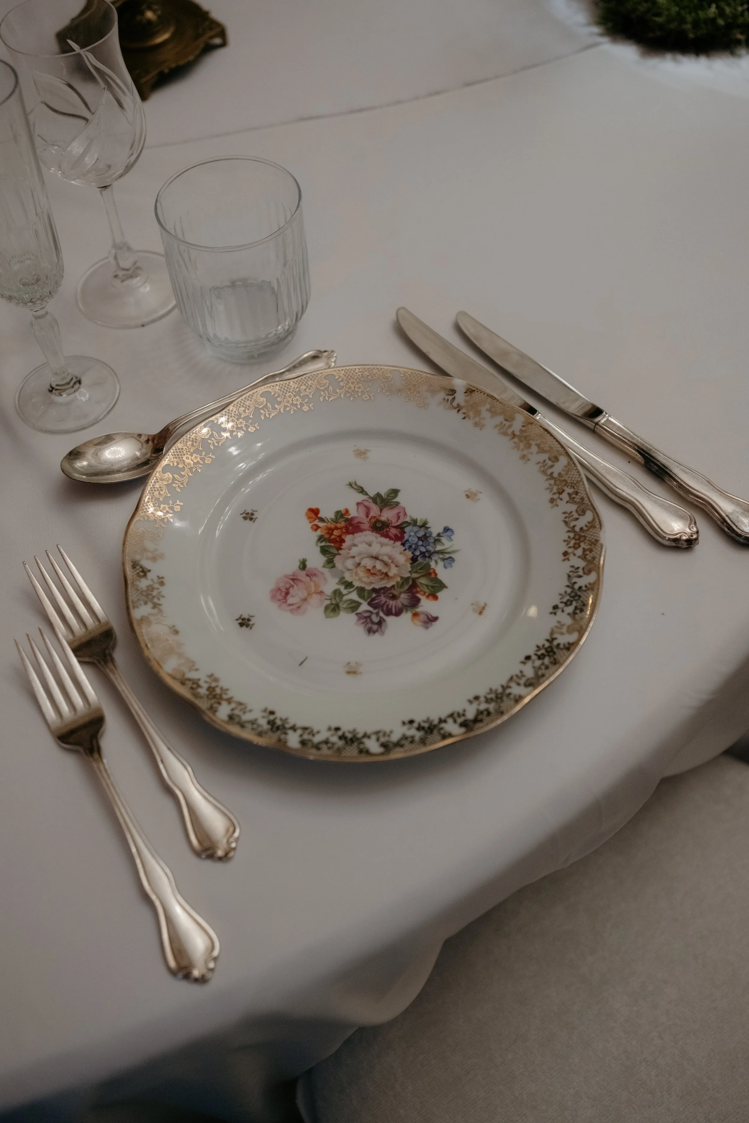 Une table élégante avec une assiette en porcelaine décorée de fleurs et de motifs dorés, accompagnée de couverts en métal argenté, plusieurs verres en cristal et un verre en verre ordinaire. La nappe est blanche et la scène évoque un dîner ou un repa