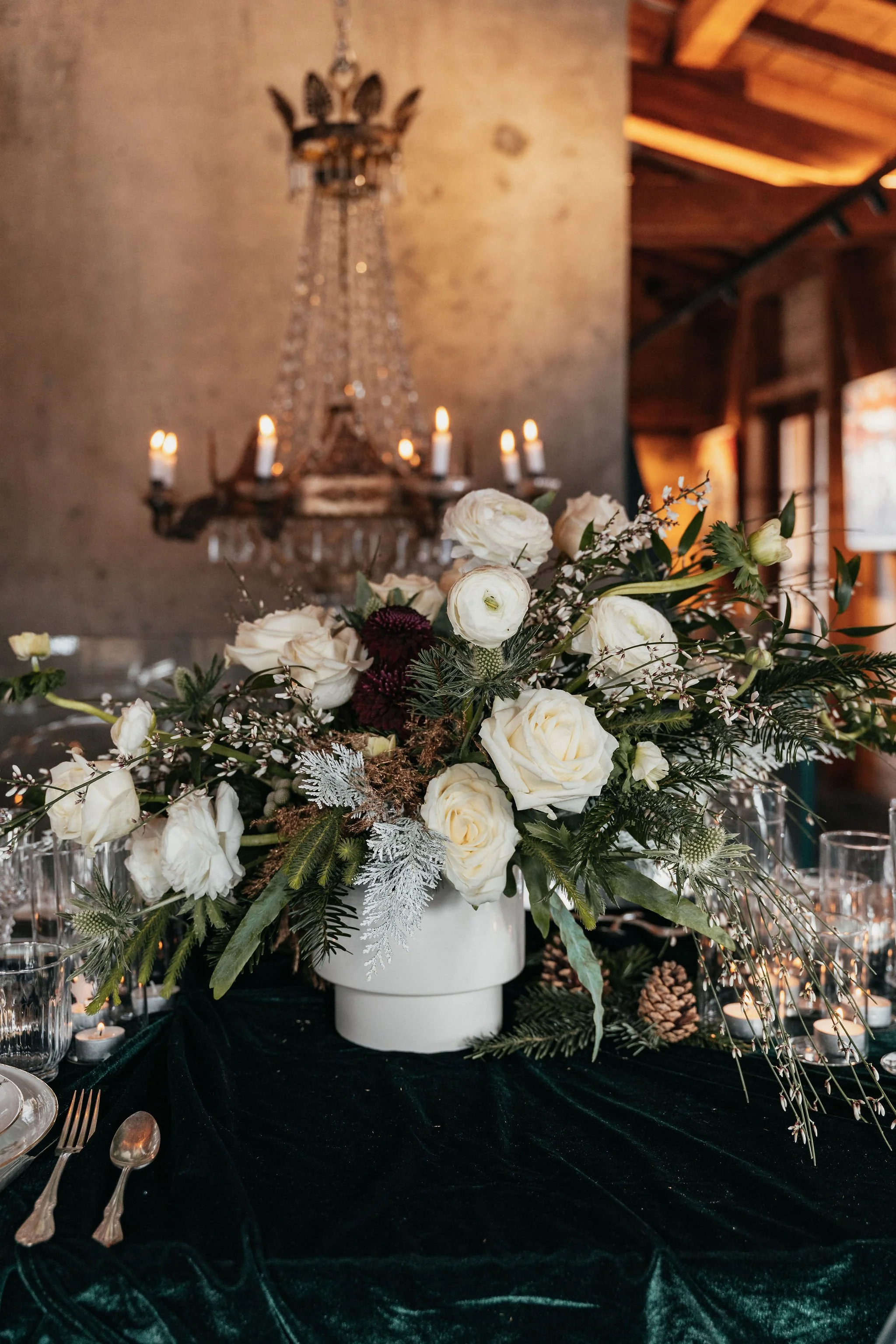 Une table décorée avec un grand bouquet de fleurs blanches dans un vase blanc, entourée de verres et de couverts, avec un lustre en cristal en arrière-plan.