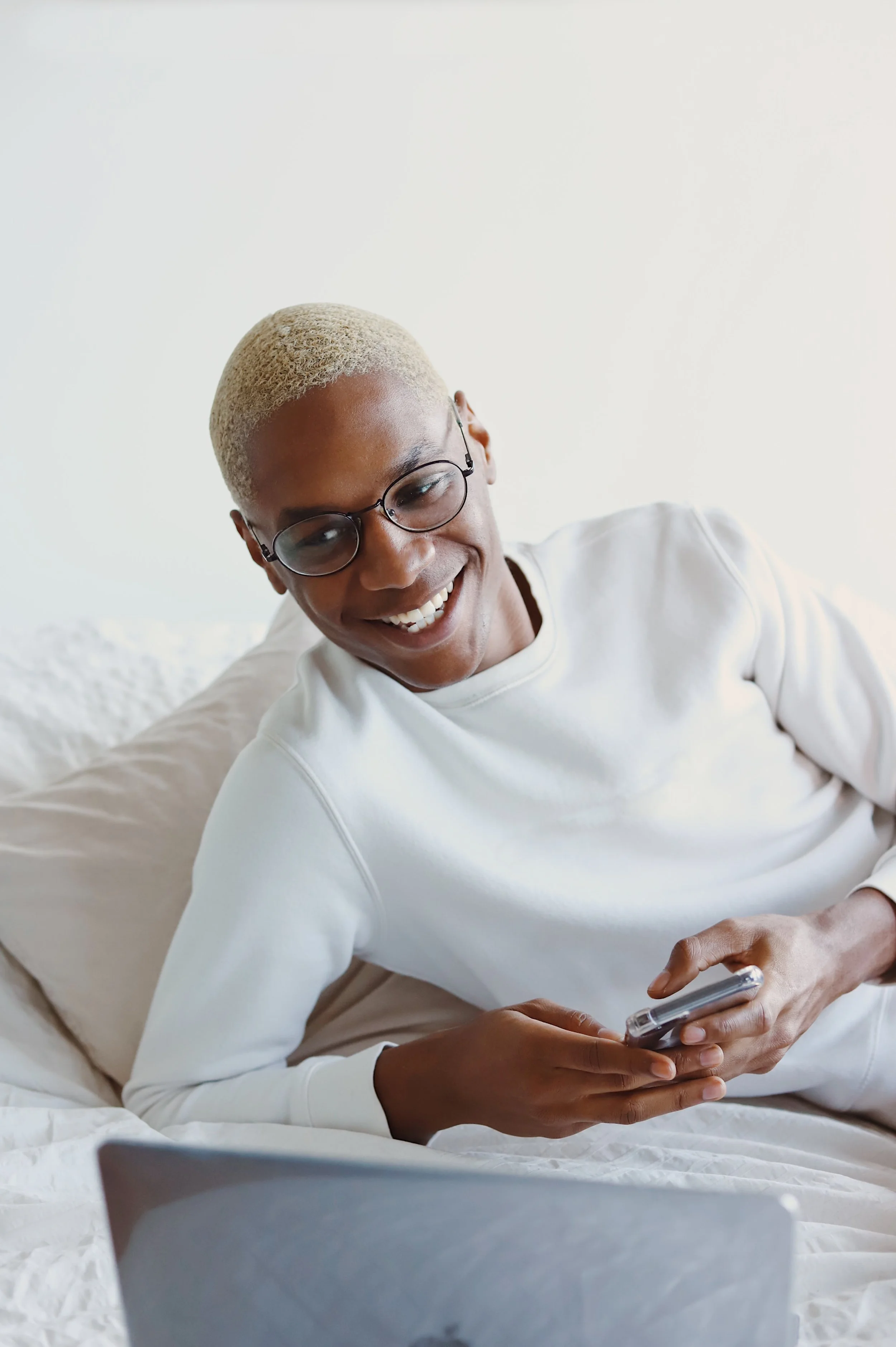 A smiling person with short blonde hair and glasses, lying on a bed with a white blanket, holding a smartphone, and looking at a laptop in front of them.