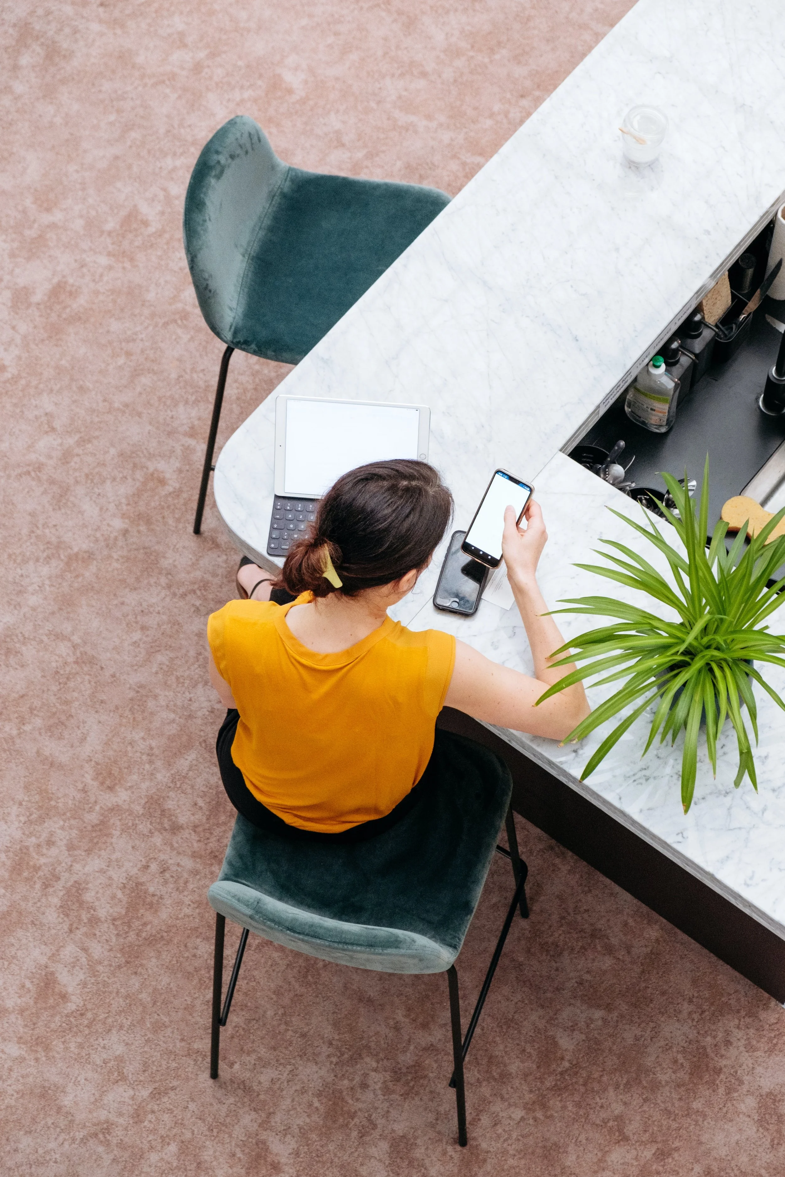 A woman sitting at a white marble countertop using her smartphone, with a laptop and green plant nearby.