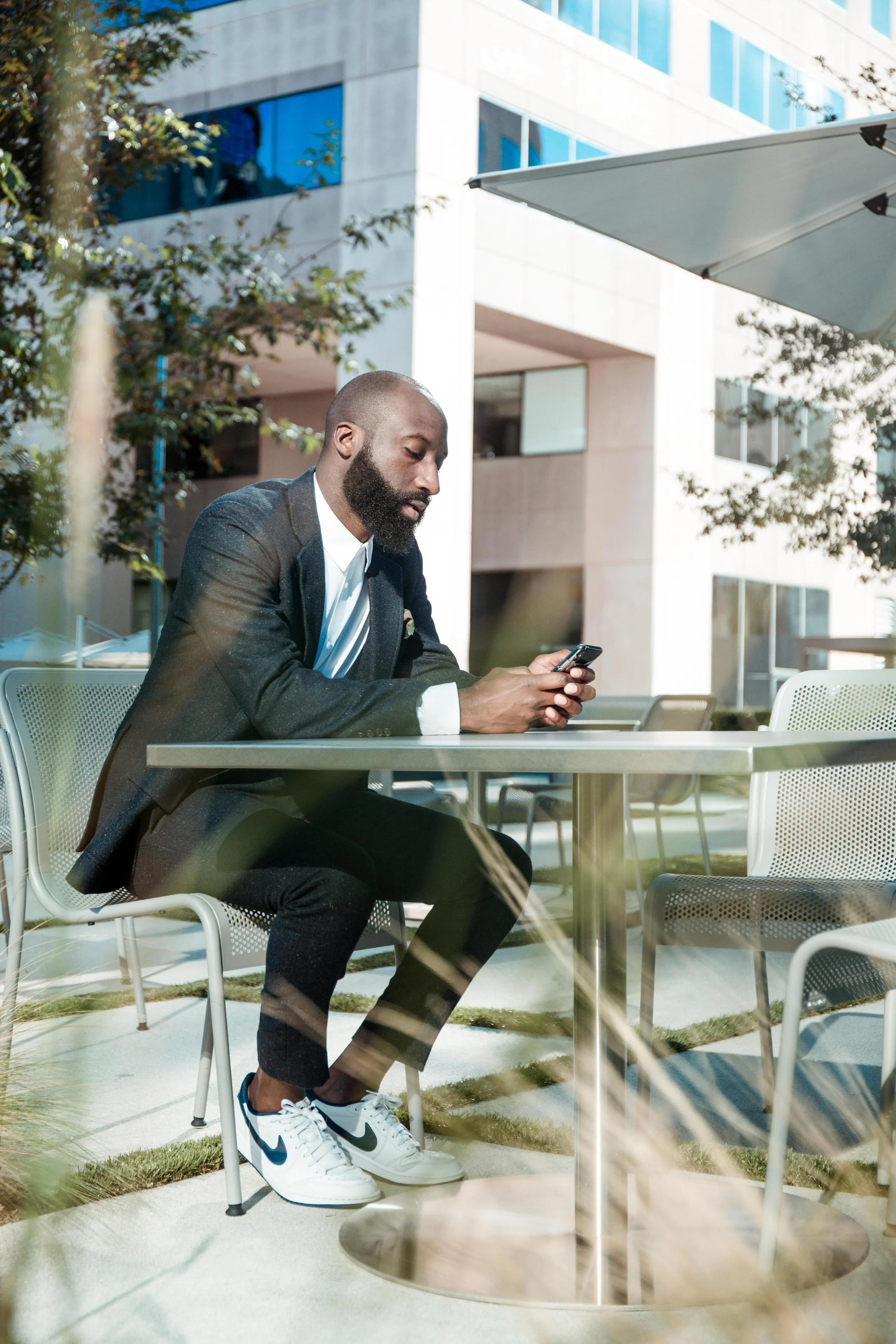 A man in a black blazer and sneakers sitting at an outdoor table using his smartphone, with modern glass buildings and trees in the background.