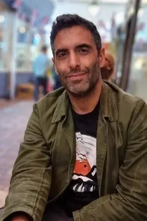Man with short dark hair and a beard, wearing a green jacket over a black T-shirt, sitting outdoors with blurred background.