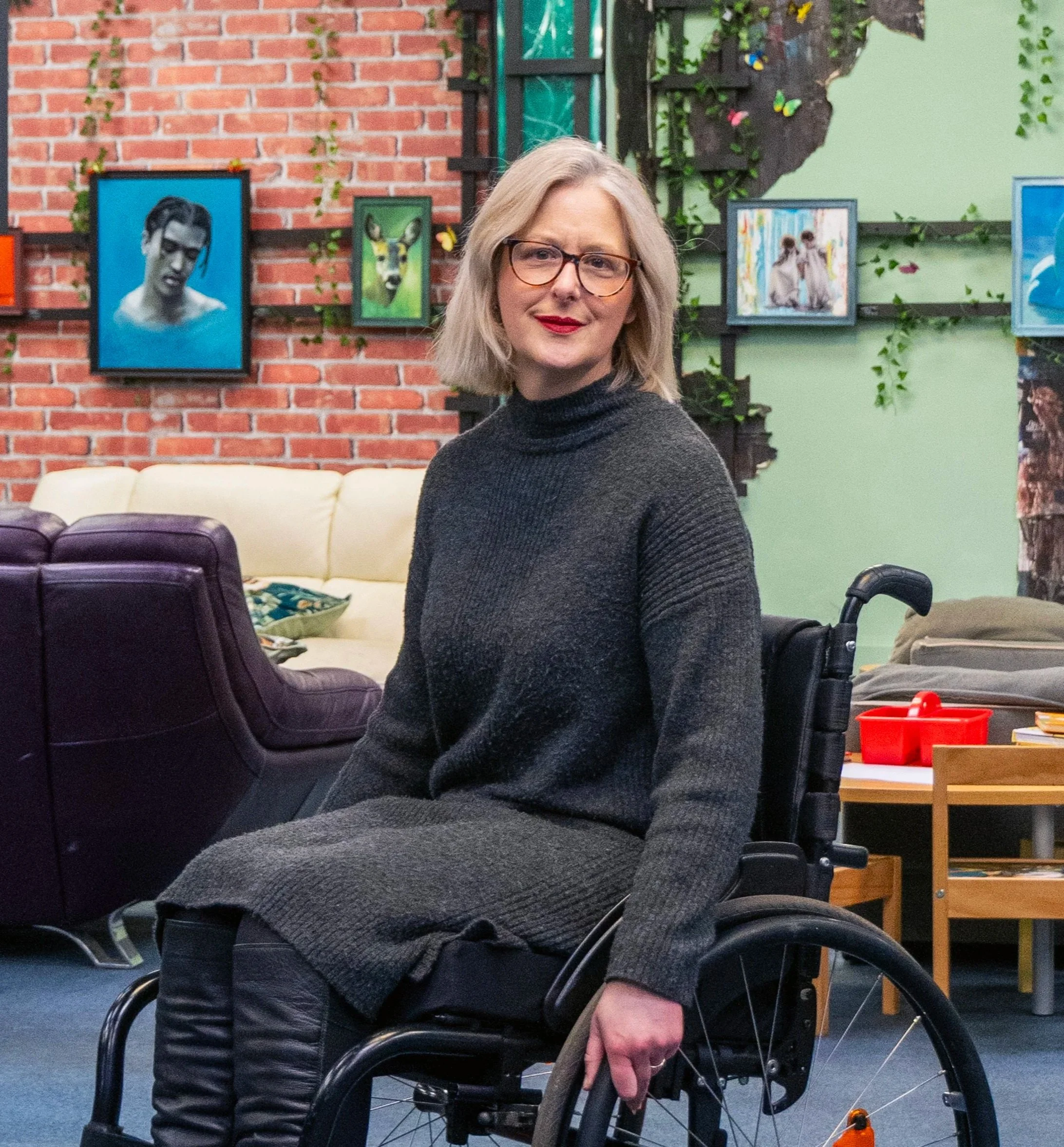 A woman with grey hair, glasses, wearing a dark grey sweater-dress, sitting in a wheelchair in a colourful room with art on the walls.