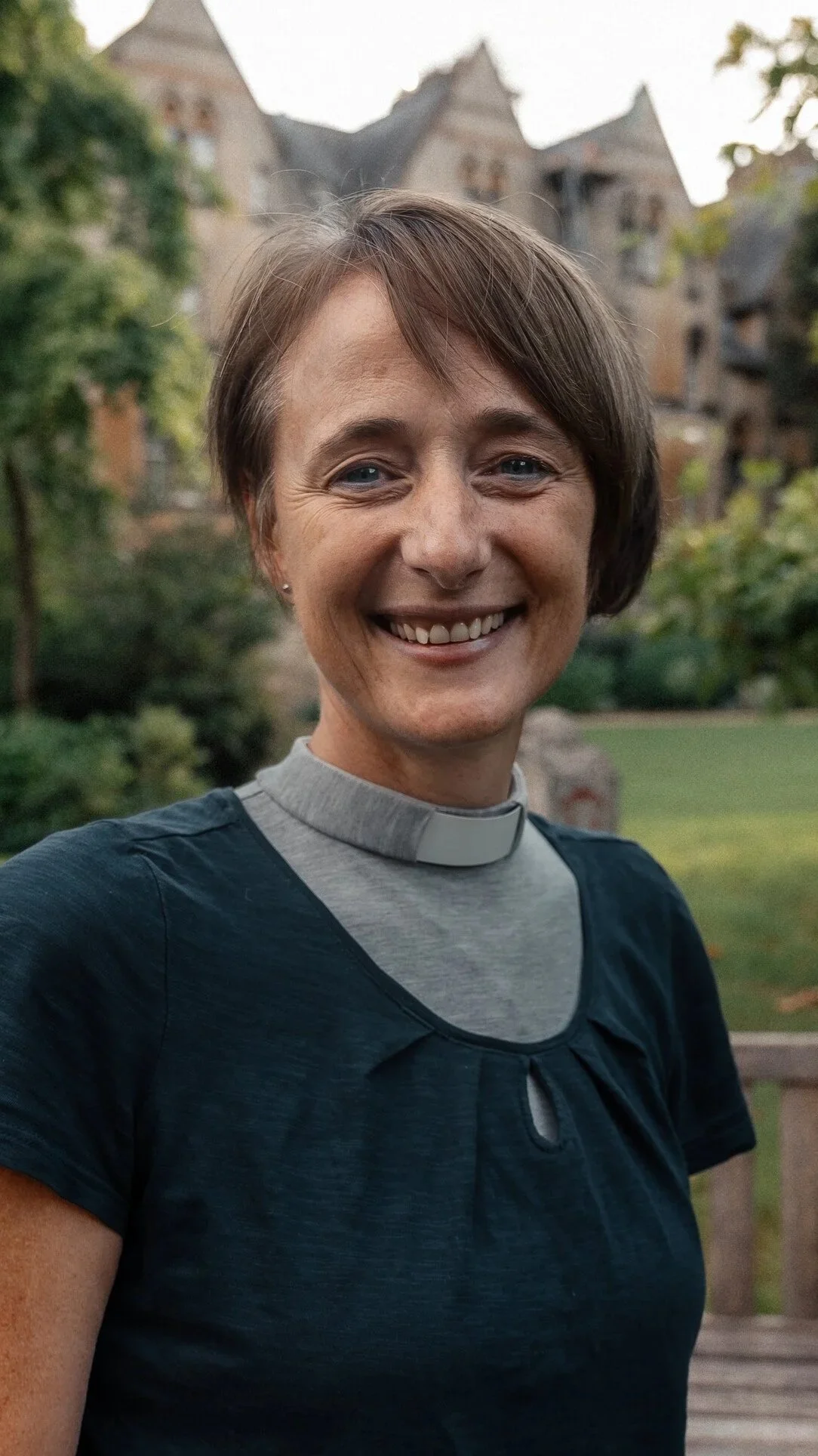 A smiling woman in clergy attire, outdoors with a historic Oxford building in the background.