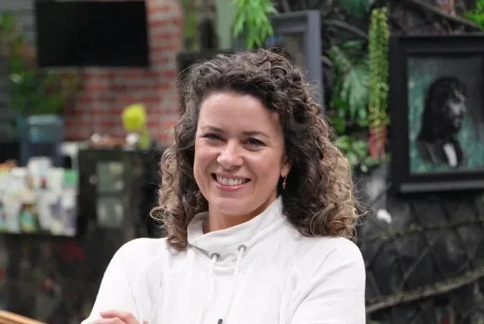 A woman with curly brown hair smiling, wearing a white coat, standing outdoors with a blurred background including plants and artwork.