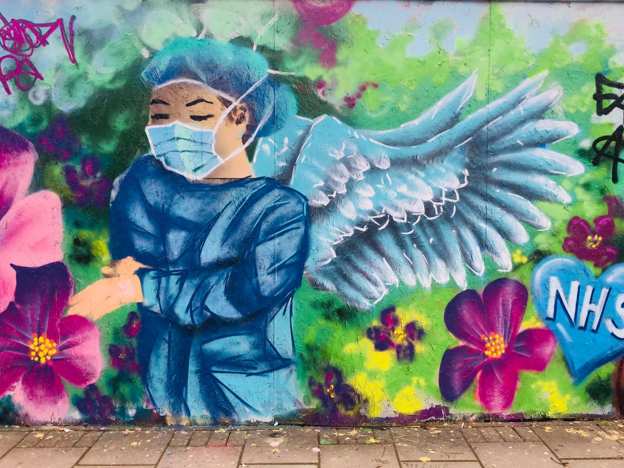 Street art mural of a person with blue hair wearing a face mask, surrounded by colorful flowers and greenery, with white angel wings on their back.