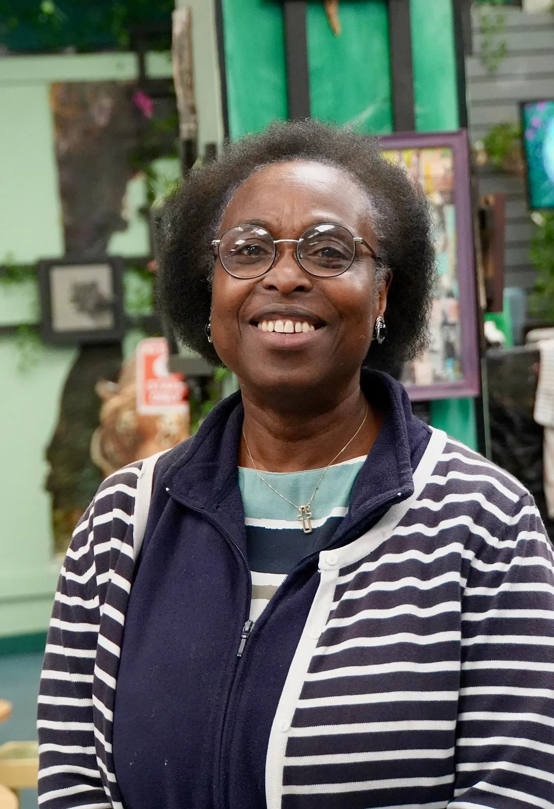 A smiling woman with short curly hair and glasses, wearing a striped shirt and a navy vest, standing indoors in front of a background with framed photos and artwork.