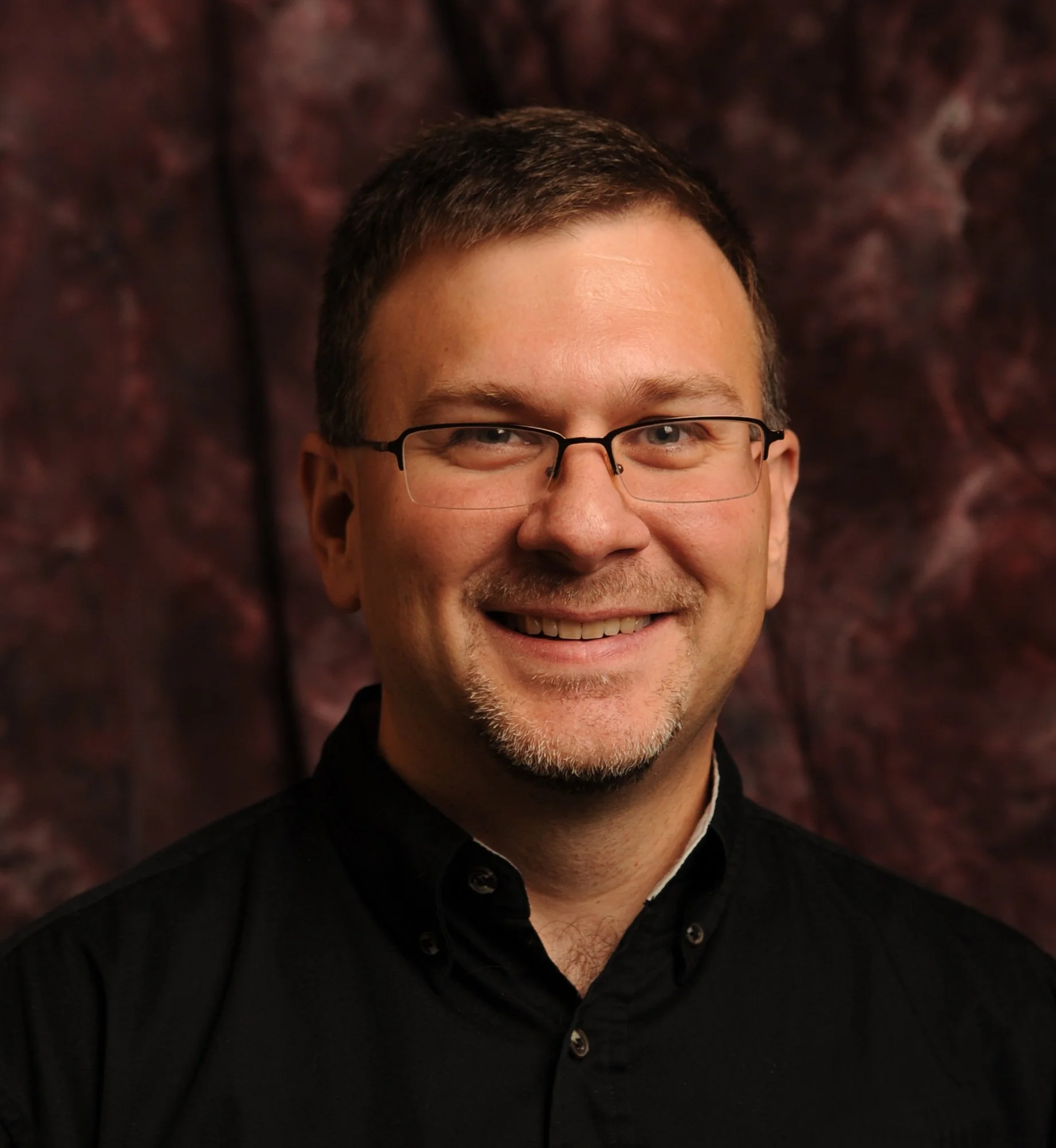 A smiling man with short brown hair and glasses, wearing a black shirt, against a dark, textured background.