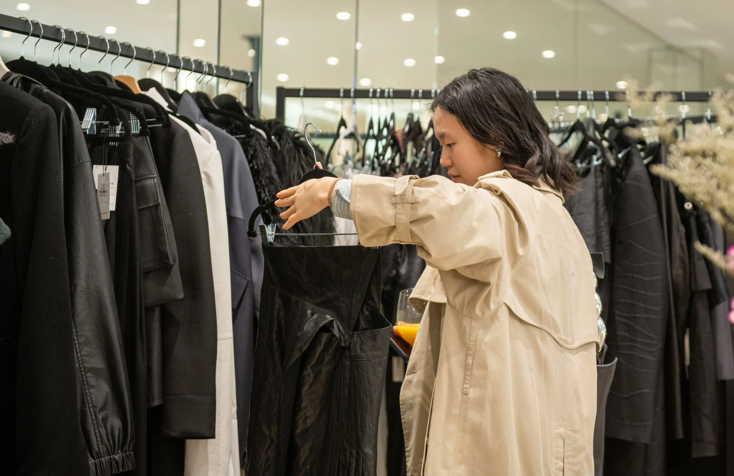 A woman shopping for black clothing at a retail store, examining a black garment on a hanger among other racks of dark-colored clothes.