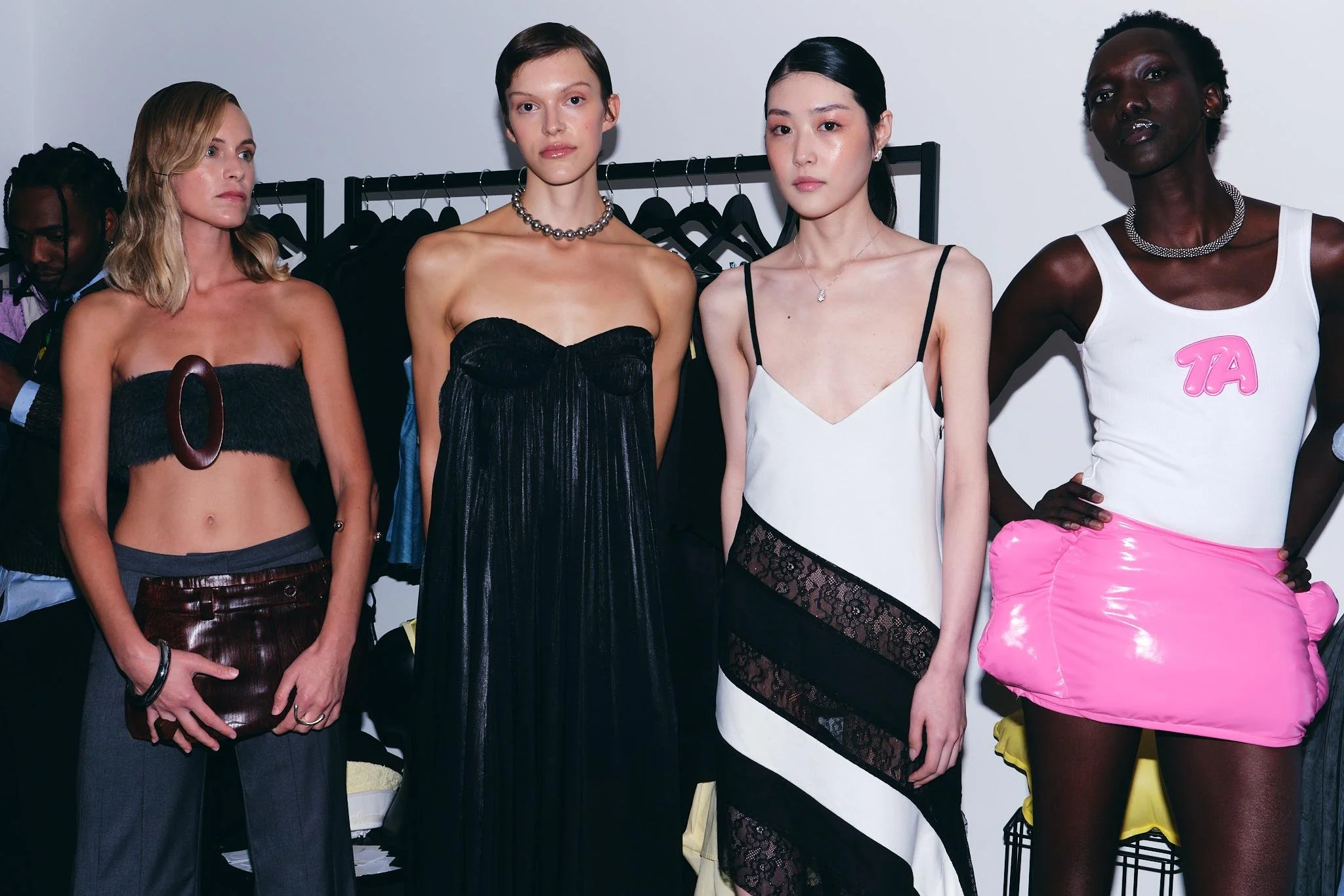 Group of five diverse women posing at a fashion event, dressed in stylish outfits with a clothing rack in the background.