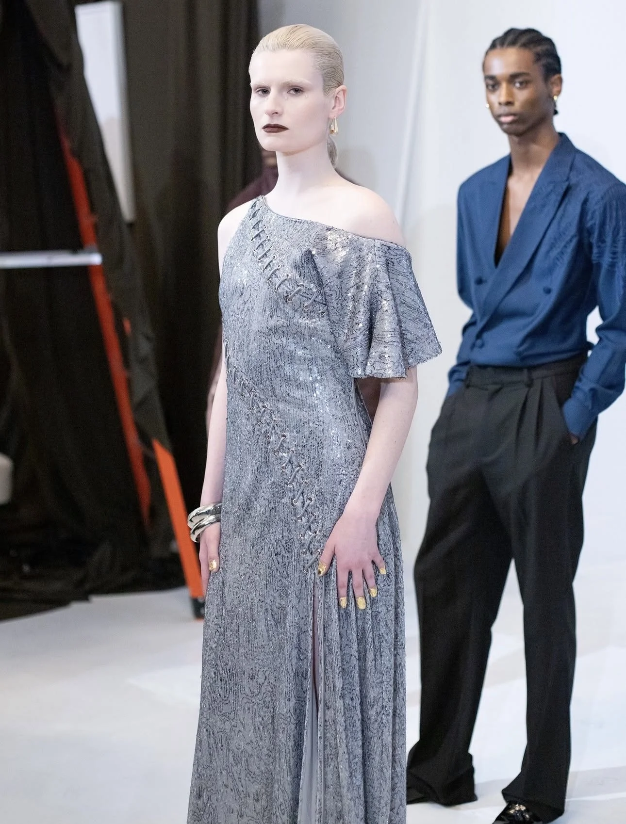 Fashion models backstage during a photoshoot, one female model in a silver, textured off-shoulder gown with dark lipstick and a male model in a blue shirt with black pants in the background.