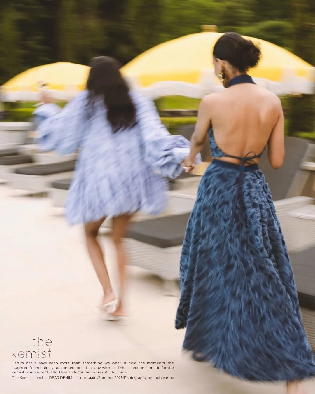 Two women, one in a blue strapless dress with an open back and the other in a long, light blue patterned dress, holding hands and running outdoors near pool loungers and yellow umbrellas.