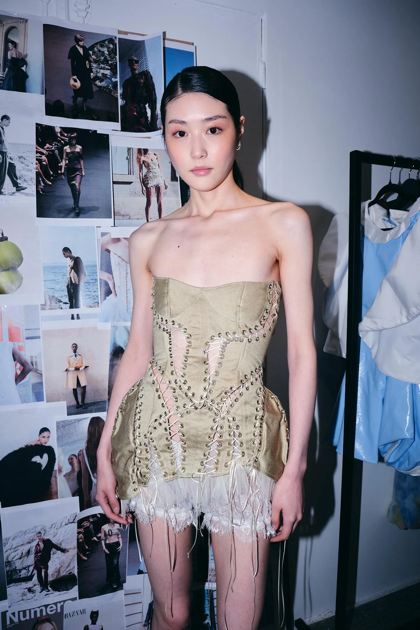 A young woman with dark hair pulled back, wearing a beige corset dress with lace and string details, standing in front of a collage of fashion photos on a wall. To her right, there is a clothing rack with some garments.