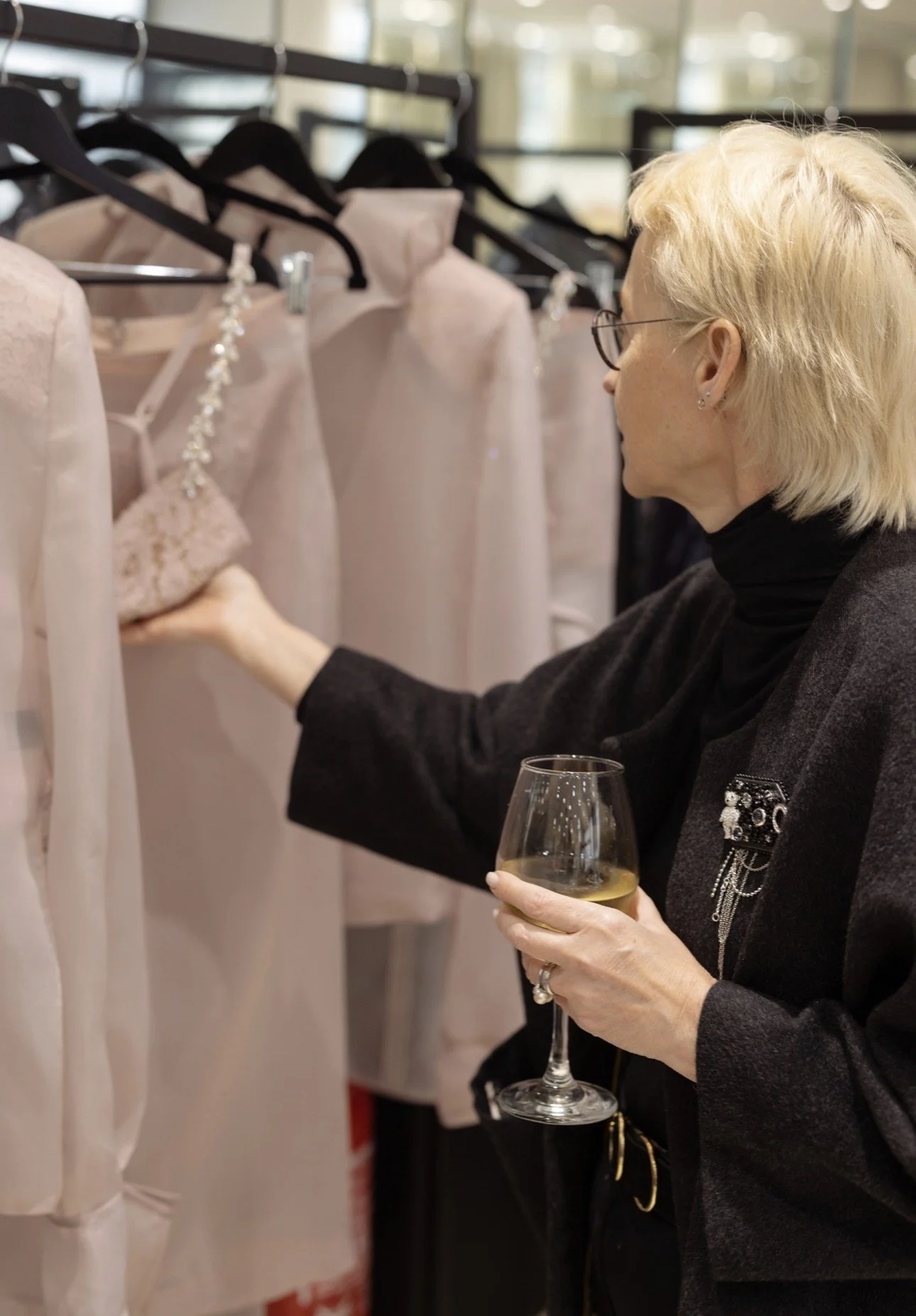 Woman with blonde hair and glasses shopping for dresses at a clothing store, holding a glass of white wine.