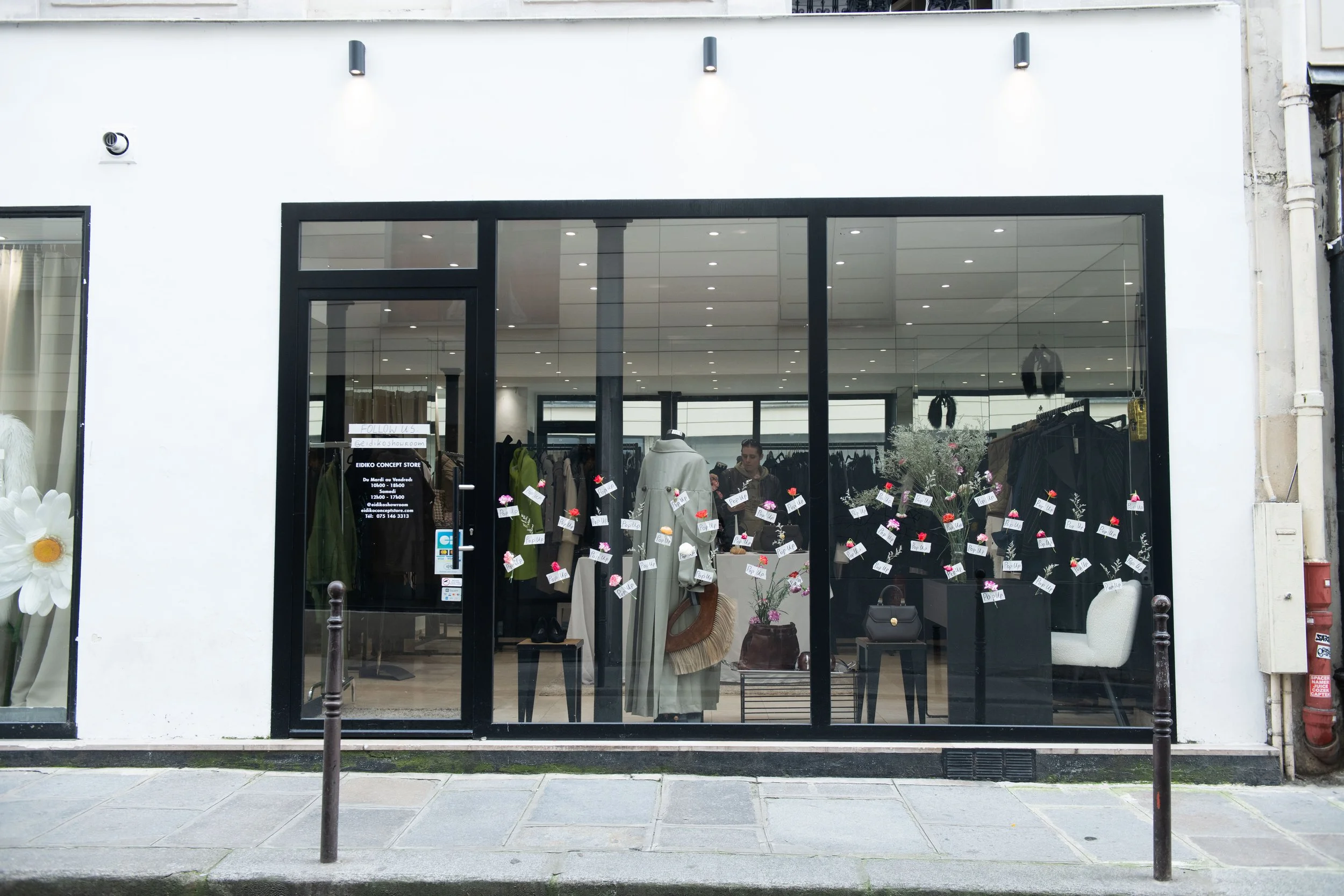 Front of a clothing store with large glass windows displaying mannequins dressed in coats and various clothing. A sign on the door provides store hours and contact information. Outside, there are flower arrangements with small signs indicating they a