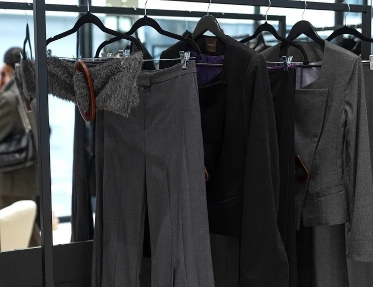 A clothing rack with gray and black women's business suits, including pants and blazers, and a gray furry accessory with brown trim hanging on a hanger.