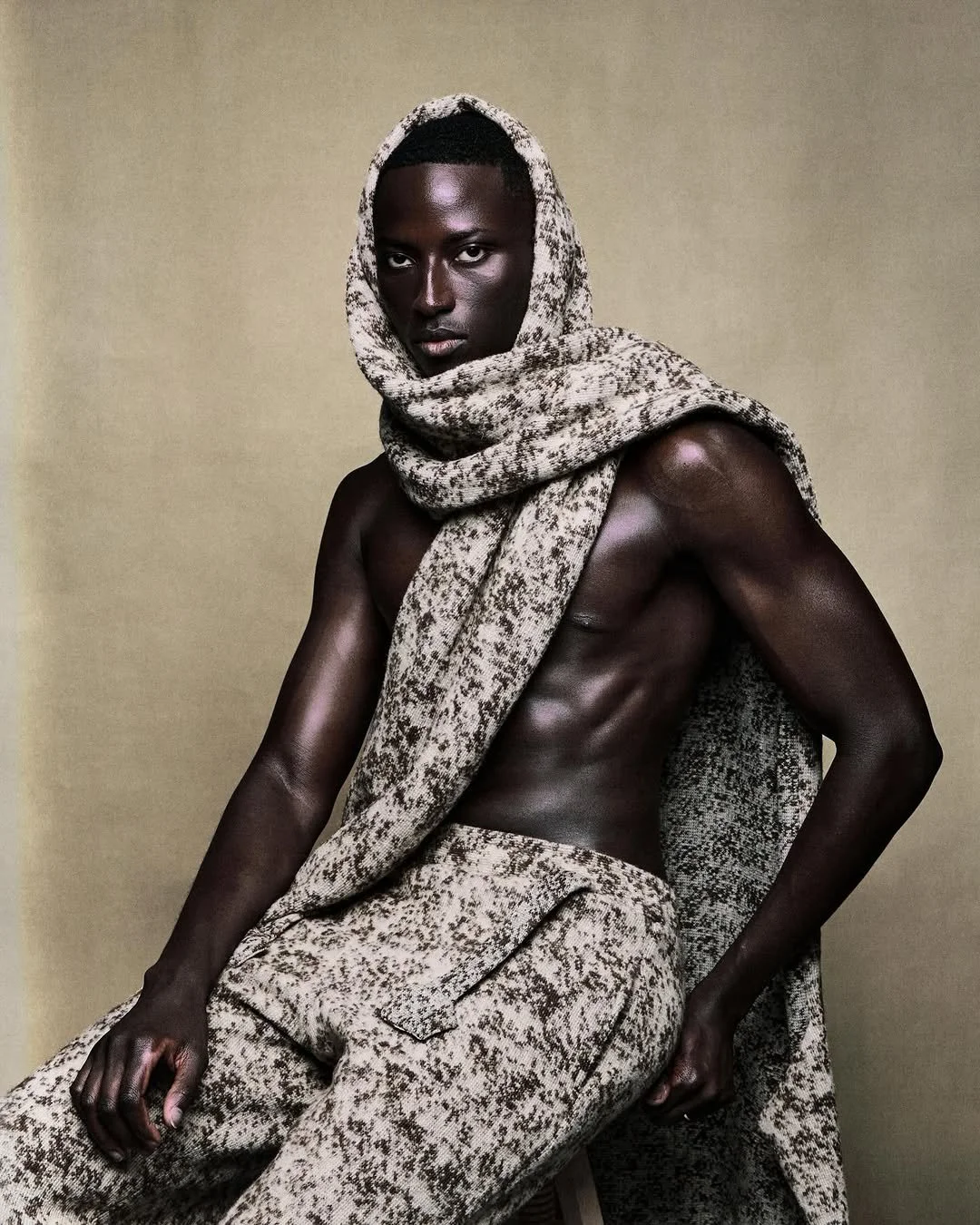 A shirtless man with dark skin, wearing snake-patterned pants and a matching scarf wrapped around his head and neck, poses against a beige background.