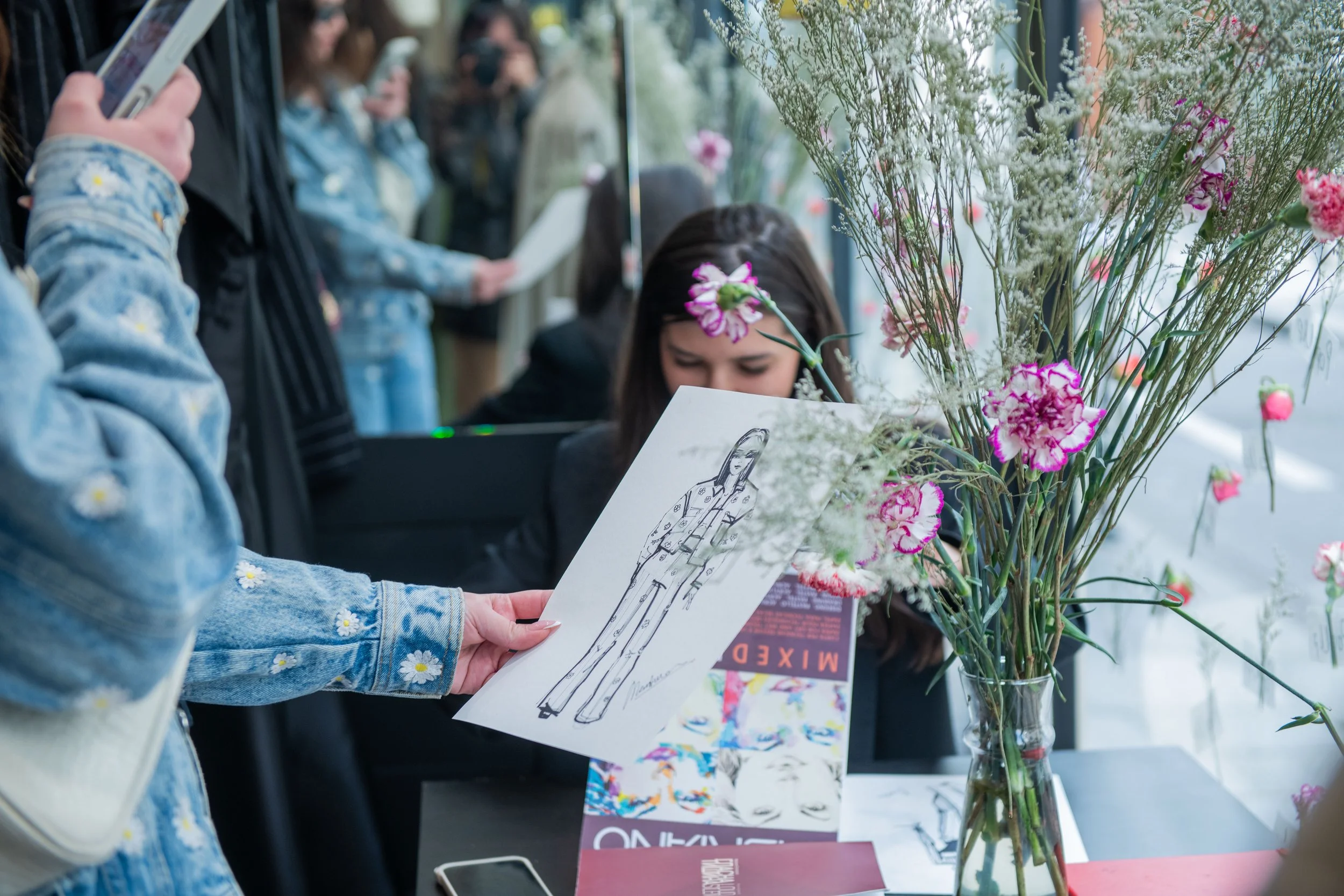 A person holding a fashion illustration in front of a display of various colorful flowers in a glass vase. In the background, other people are taking photos and reading at an event or exhibit.