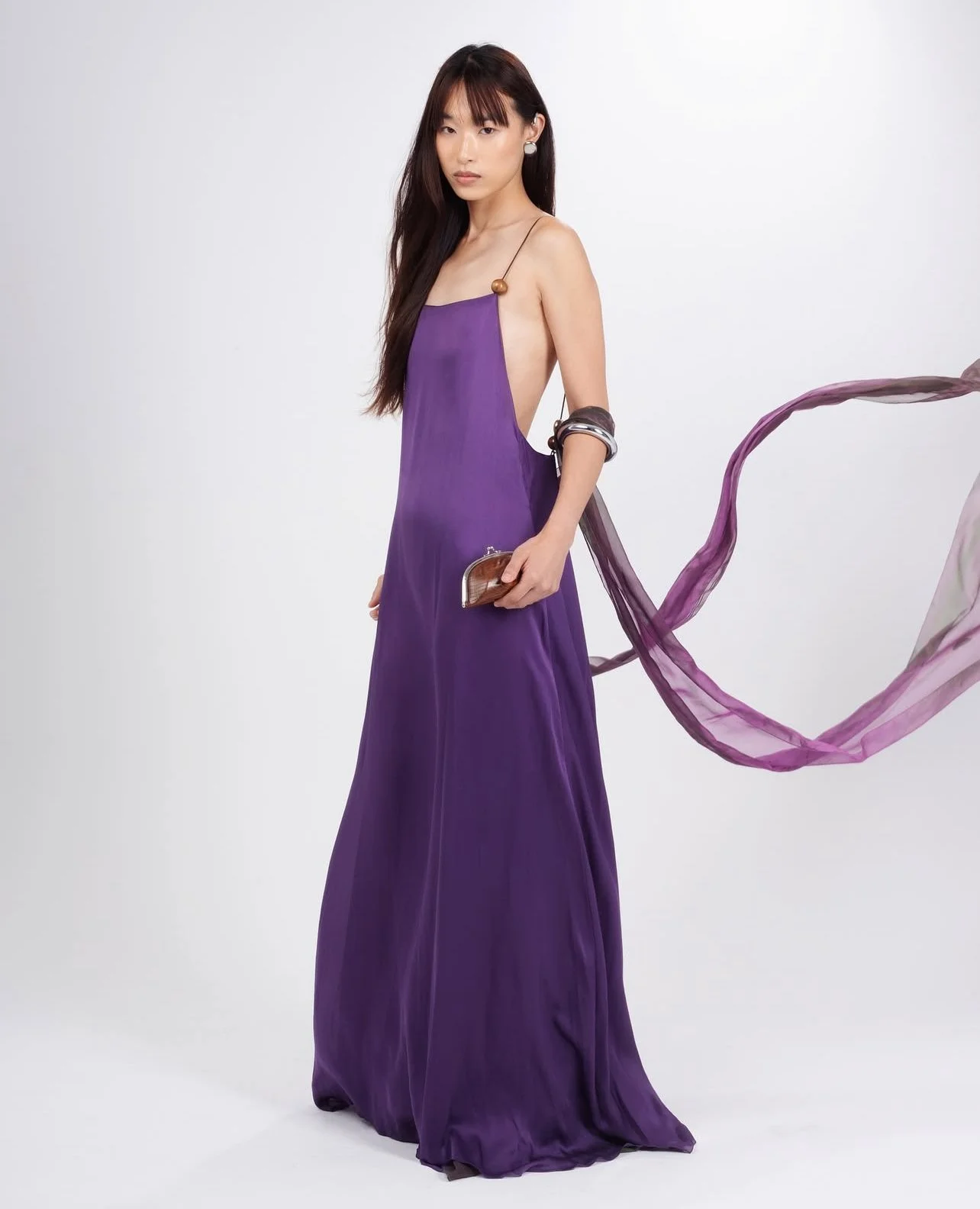 A woman in a long purple gown with side cutouts holds a clutch purse, with sheer purple fabric flowing behind her, against a plain white background.