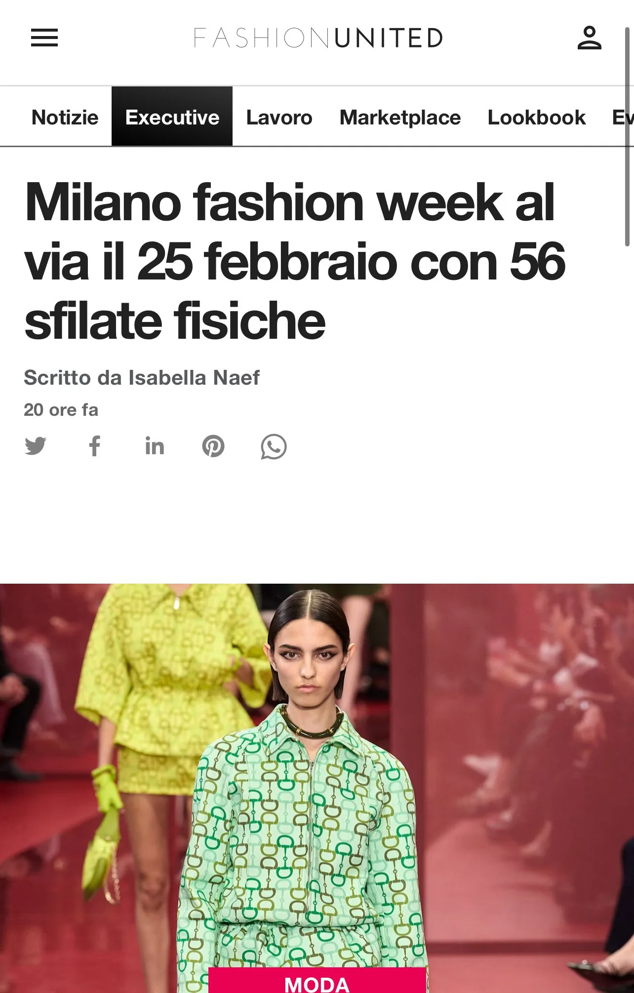 Fashion runway show with models walking in designer outfits; model in foreground wearing a patterned shirt dress with chain motif in green and cream colors, walking on a red carpet runway.