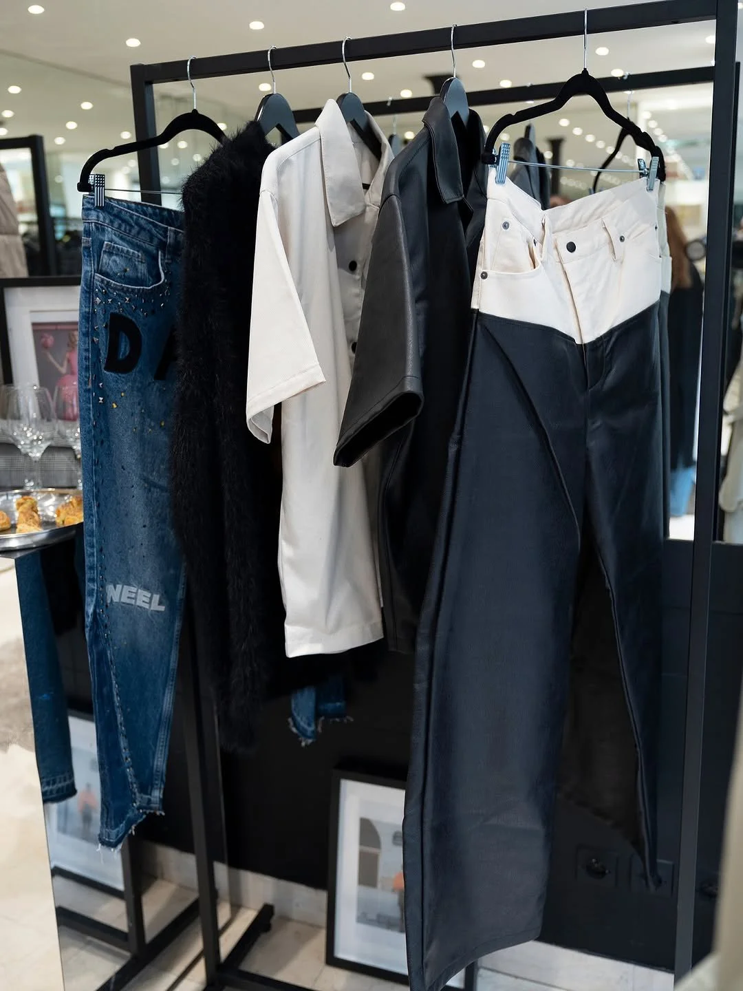 Clothing rack with black, white, and beige shirts, black pants, with a pair of jeans hanging on the left, and frames on the floor beneath. The background shows a shopping mall interior.