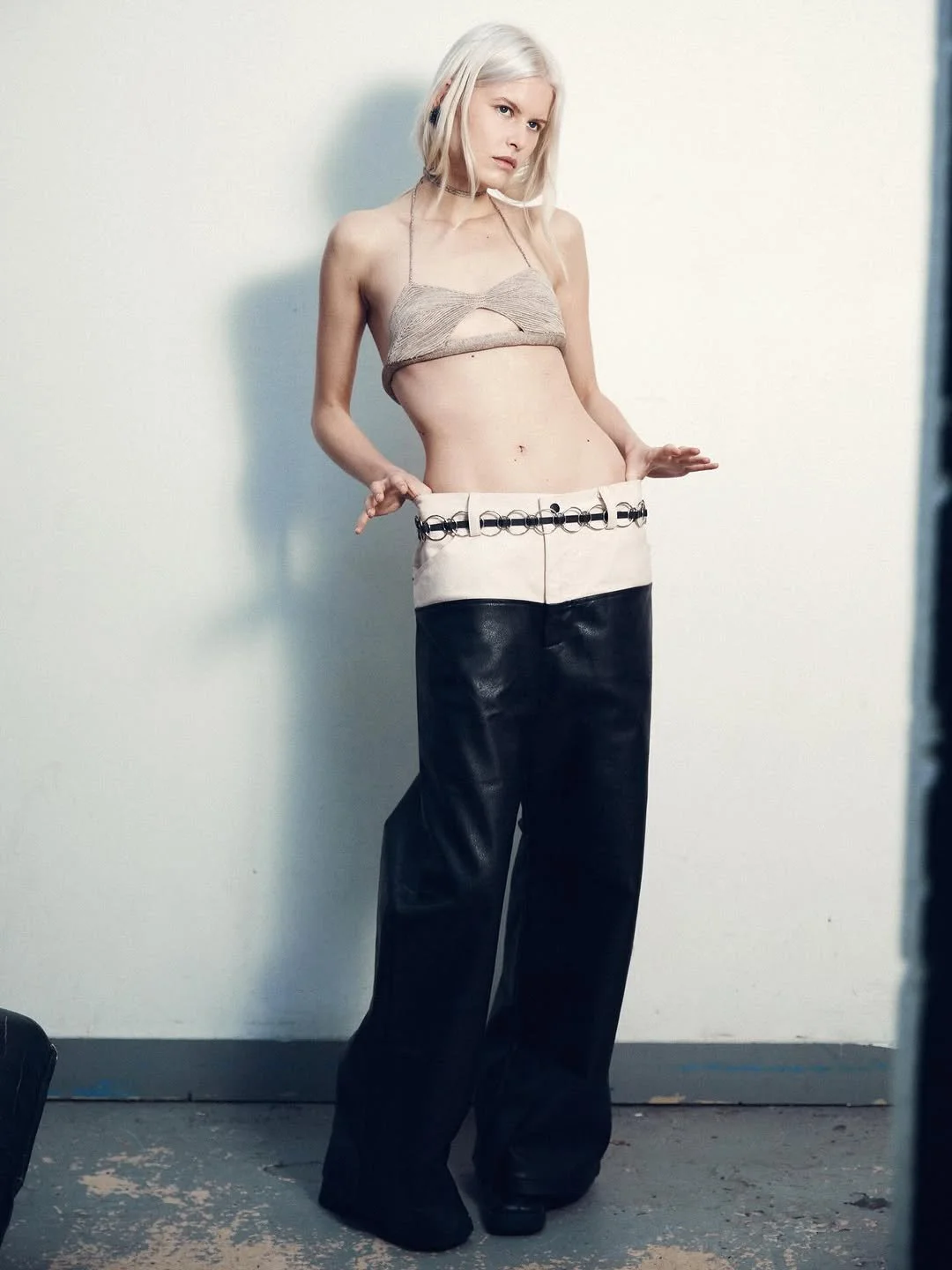 A young woman with platinum blonde hair wearing a beige spaghetti strap crop top, oversized black and white pants with metal rings, standing against a plain white wall, looking to the side with a serious expression.