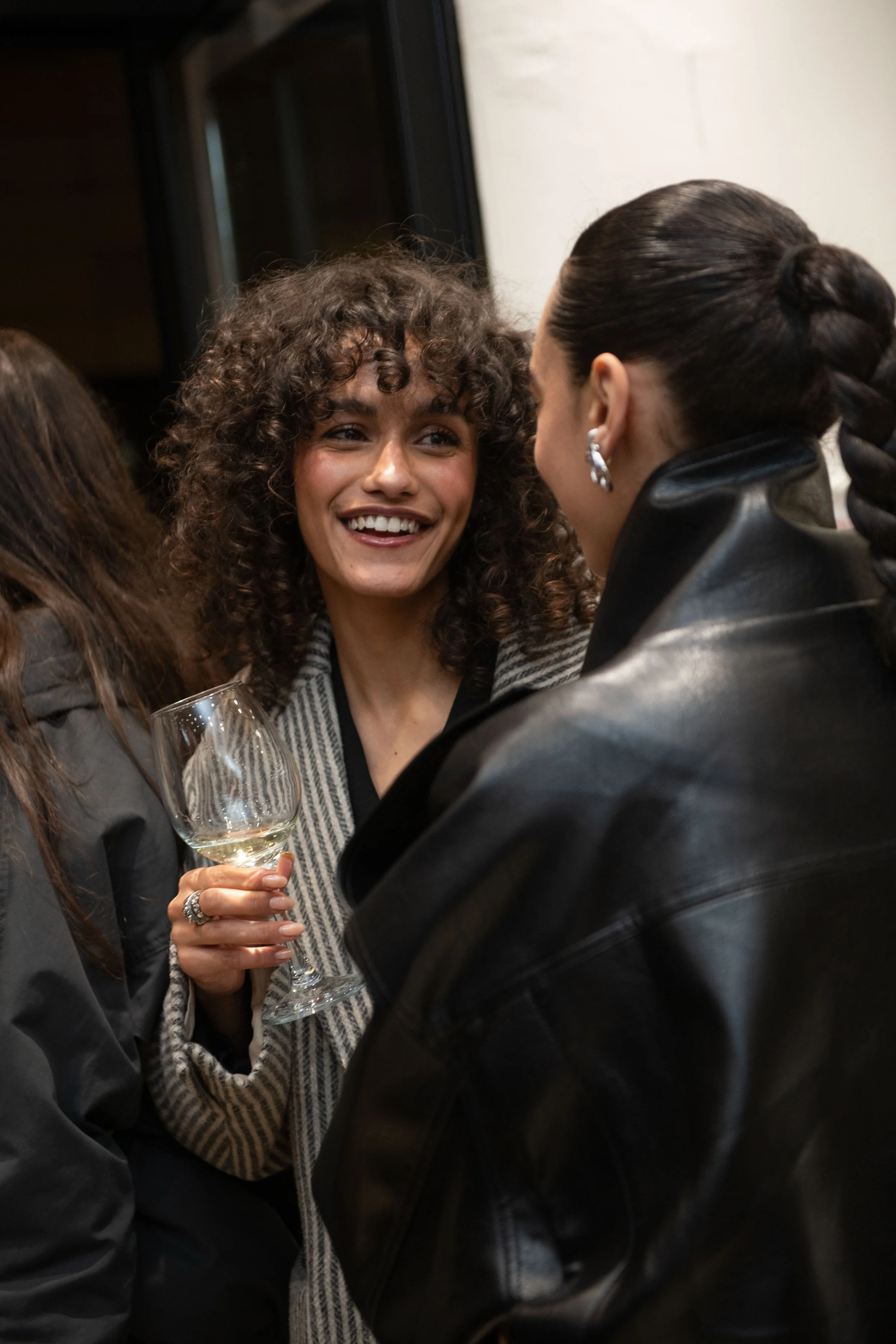 Two women are talking close to each other at a social gathering, one holding a stemmed wine glass. The woman with curly hair is smiling and wearing a striped blazer, while the other has dark hair pulled back and is wearing earrings and a black leathe