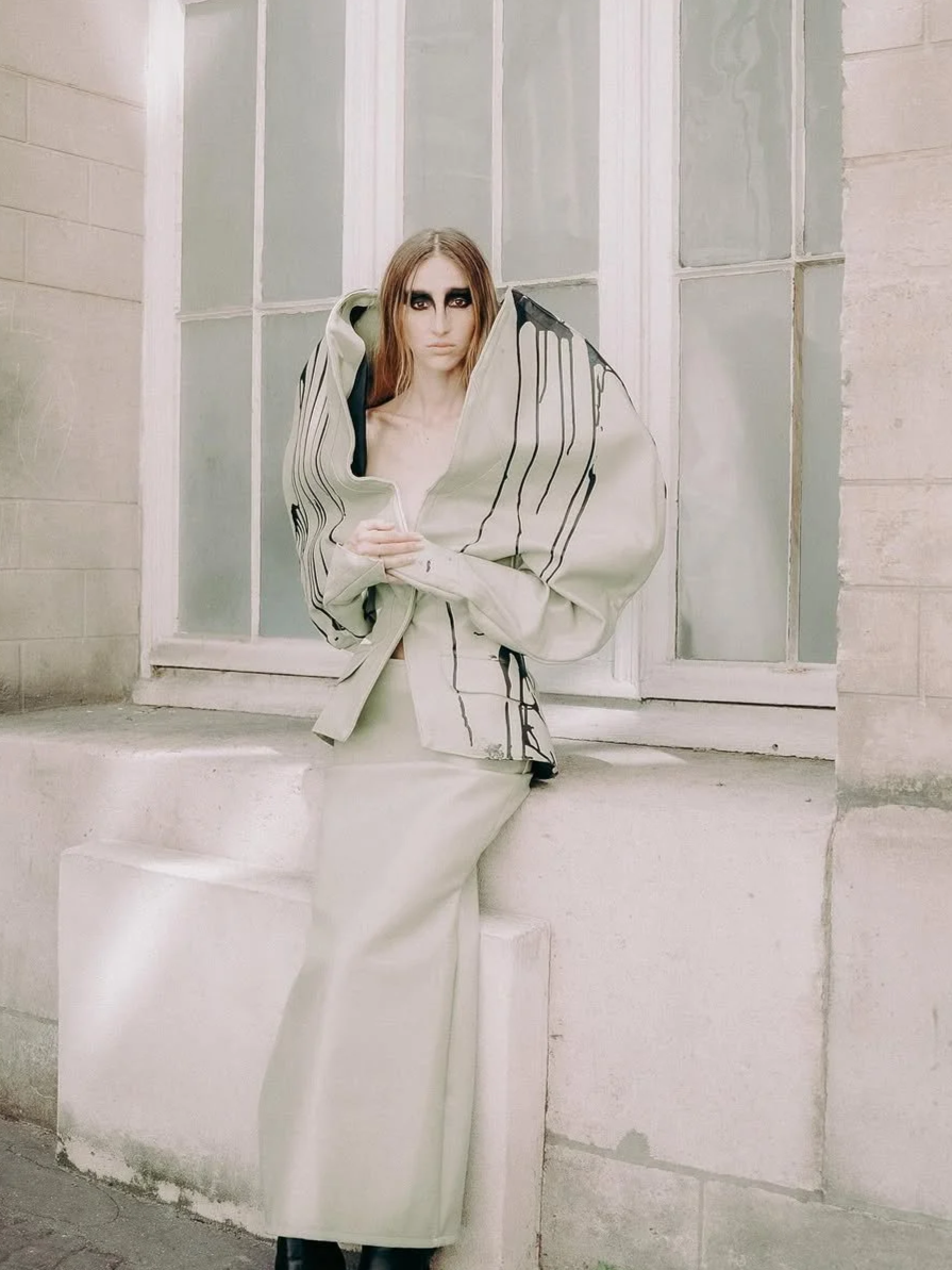 Woman dressed in beige oversized jacket and long skirt with black paint-like streaks, standing by a window with her arms crossed, against a stone building background.