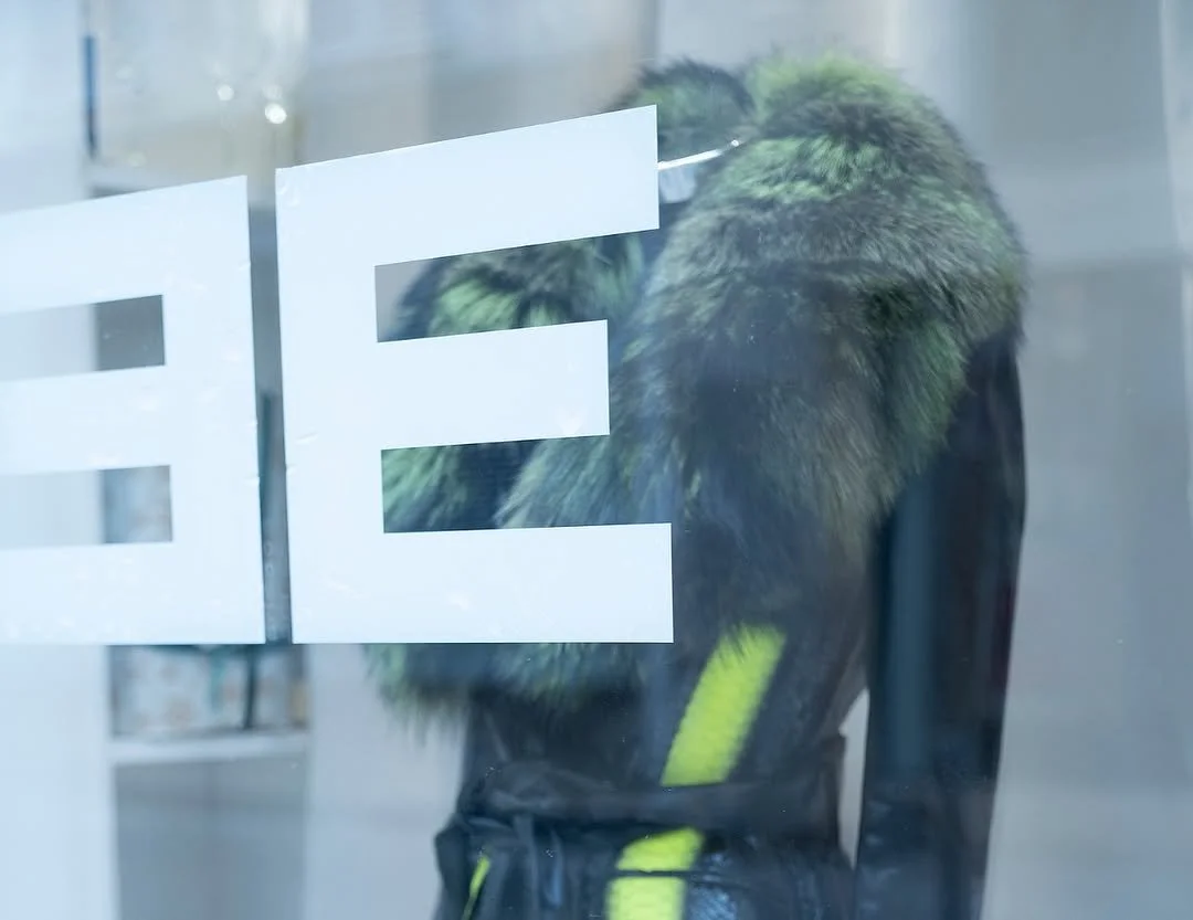 A mannequin wearing a furry green and black jacket with yellow accents, seen through a glass window with the word 'FE' partially visible.