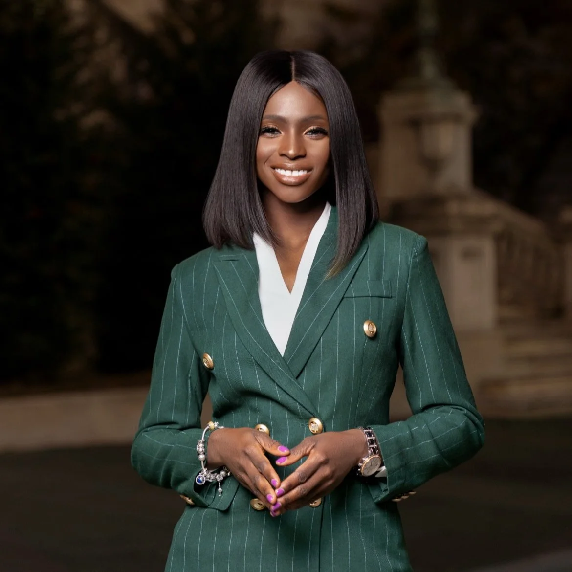 A woman wearing a green pinstripe blazer with gold buttons, standing outdoors, smiling, with dark hair styled in a straight bob.