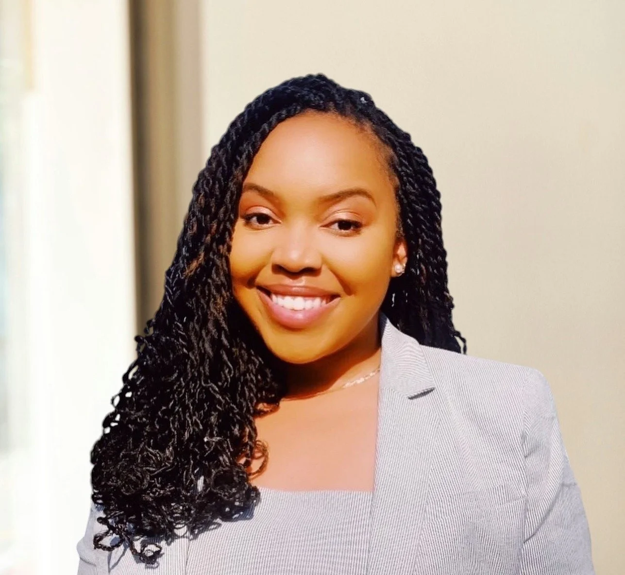 A smiling woman in a gray blazer with dark curly hair styled to one side, standing against a plain light-colored background.
