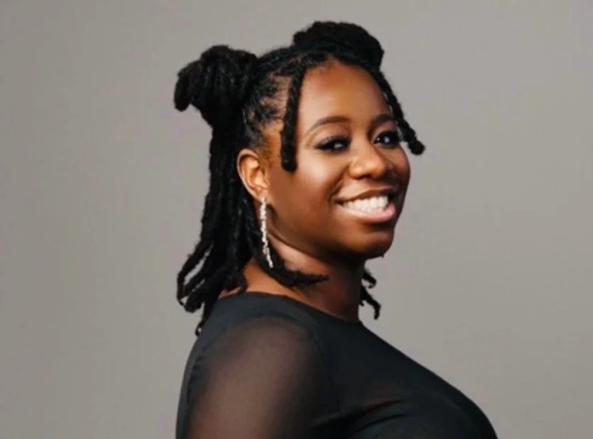 A woman with braided hair styled into two buns, wearing a black top and earrings, smiling against a plain gray background.
