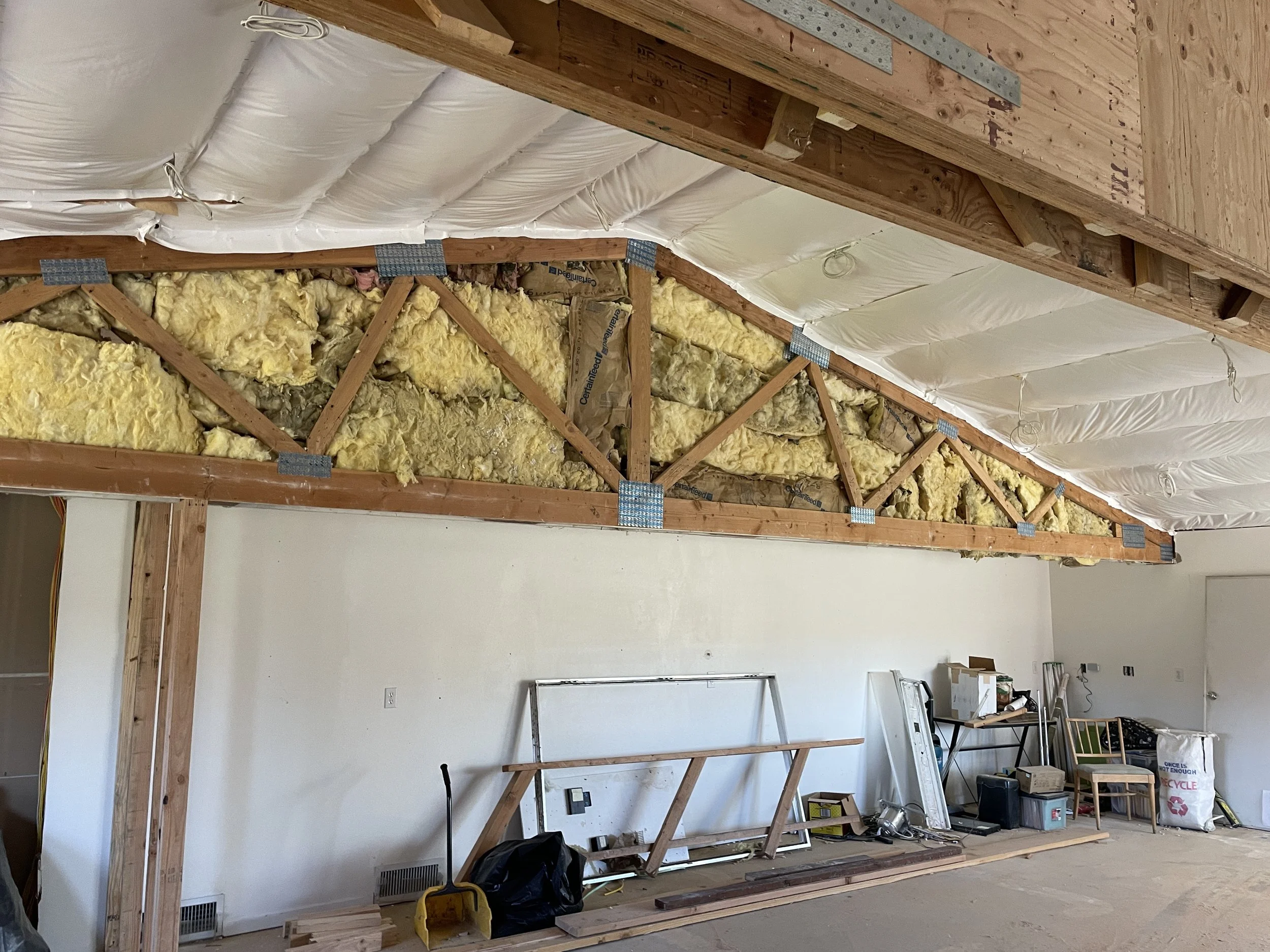 Interior view of a construction site showing unfinished walls with wood framing and insulation. Various tools and materials are scattered around, and a table is visible against the wall.
