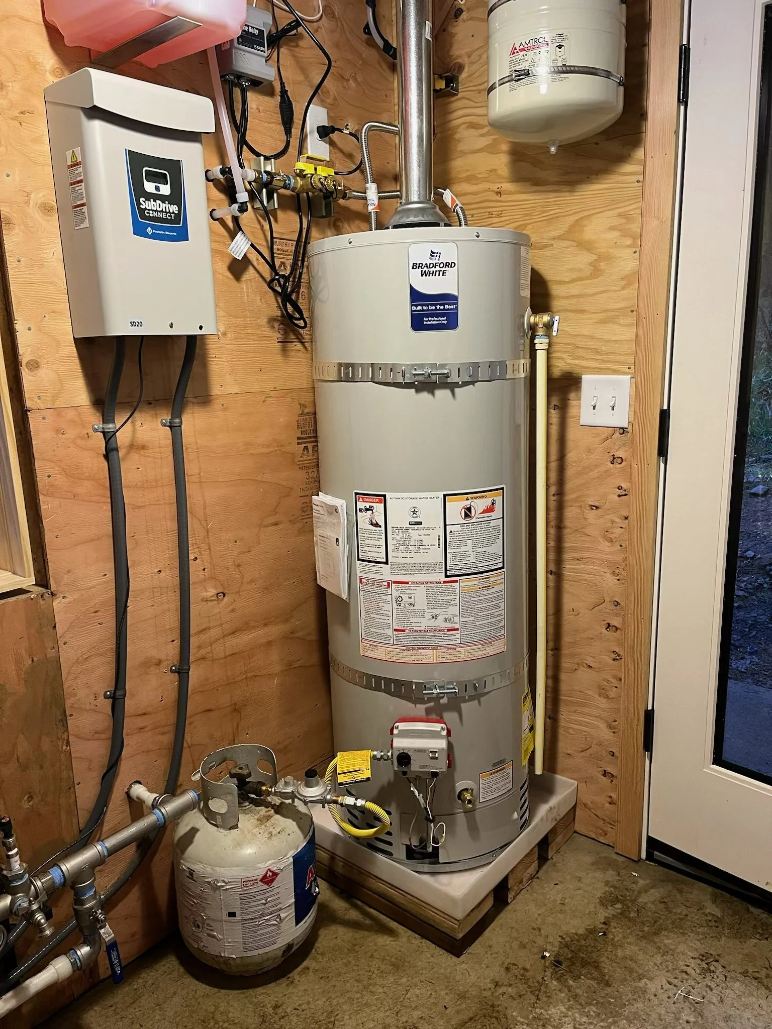 A water heater in a wooden utility room with plumbing and electrical components.