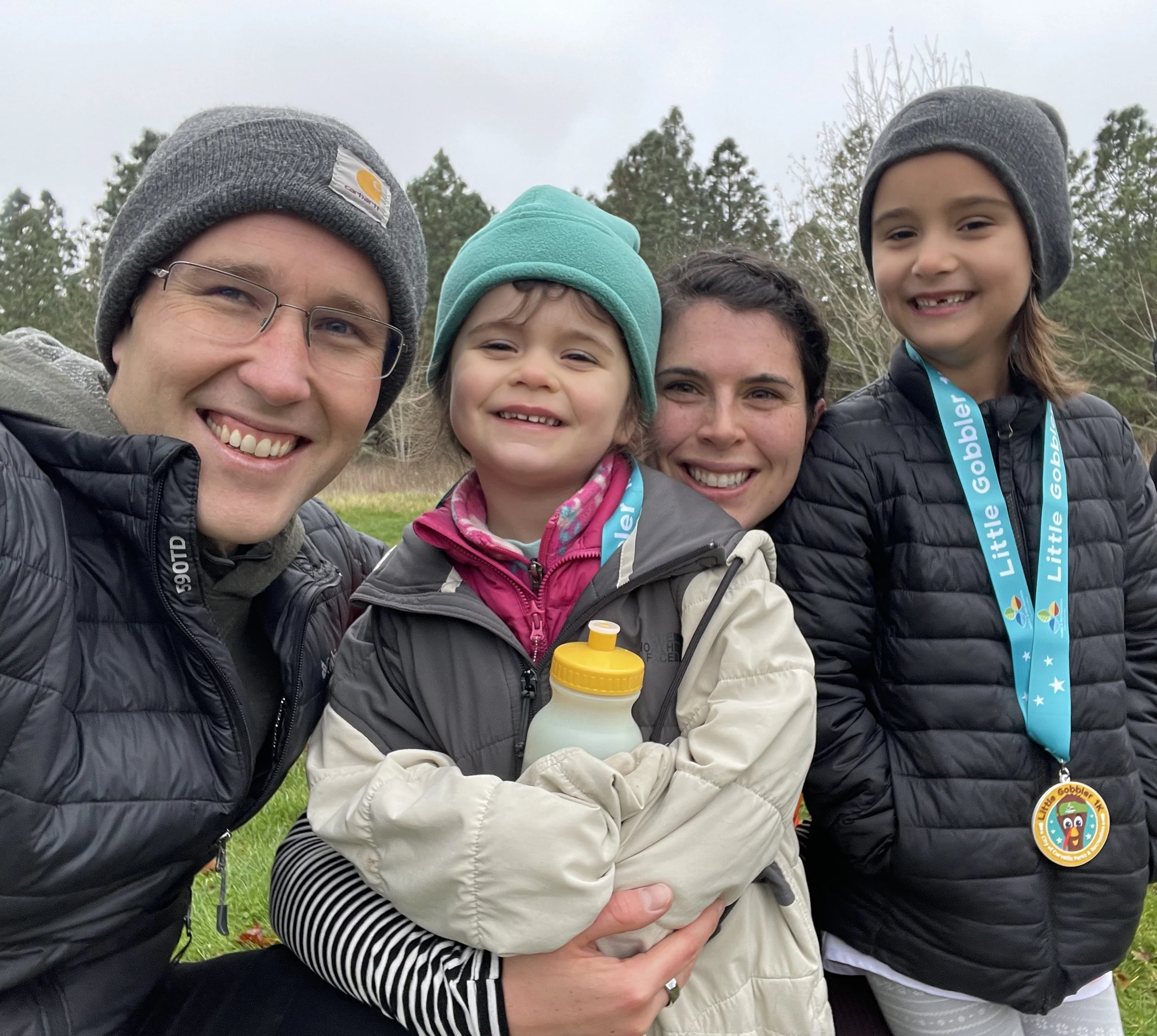A group of five people, including children and adults, smiling outdoors wearing winter clothing, with trees and an overcast sky in the background.