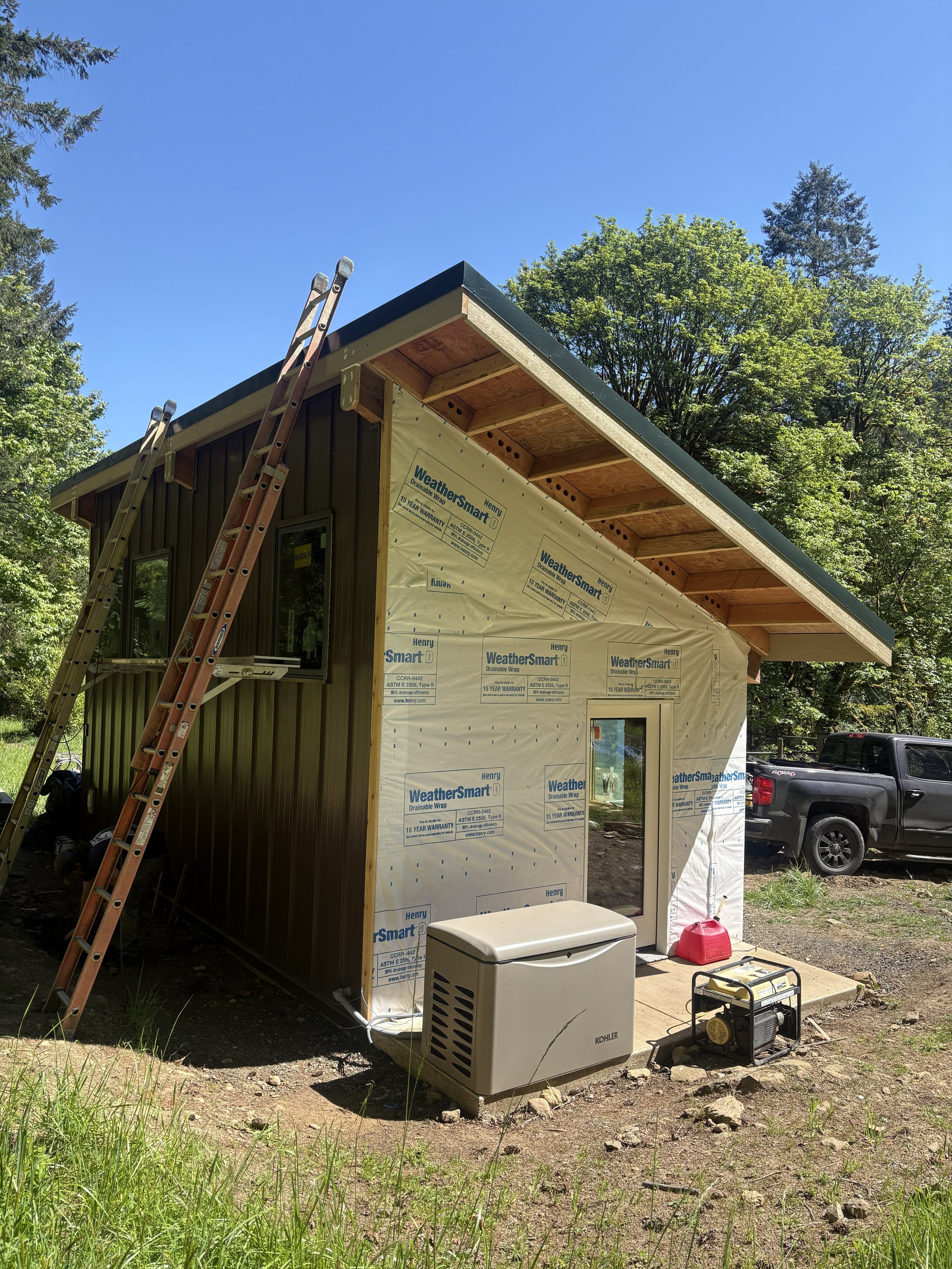 A house under construction with a partially finished exterior, an unfinished roof, and construction tools and equipment in front. The house has weather-resistant wrap on one side and a metal siding on the other. A ladder leans against the house, and 