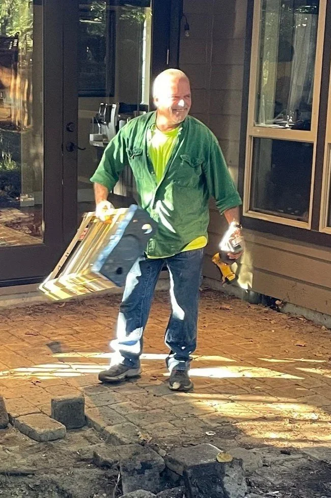 A man smiling and holding tools, including a drill and a small piece of wood, standing on a brick patio near the doorway of a house, likely engaged in a home improvement project.