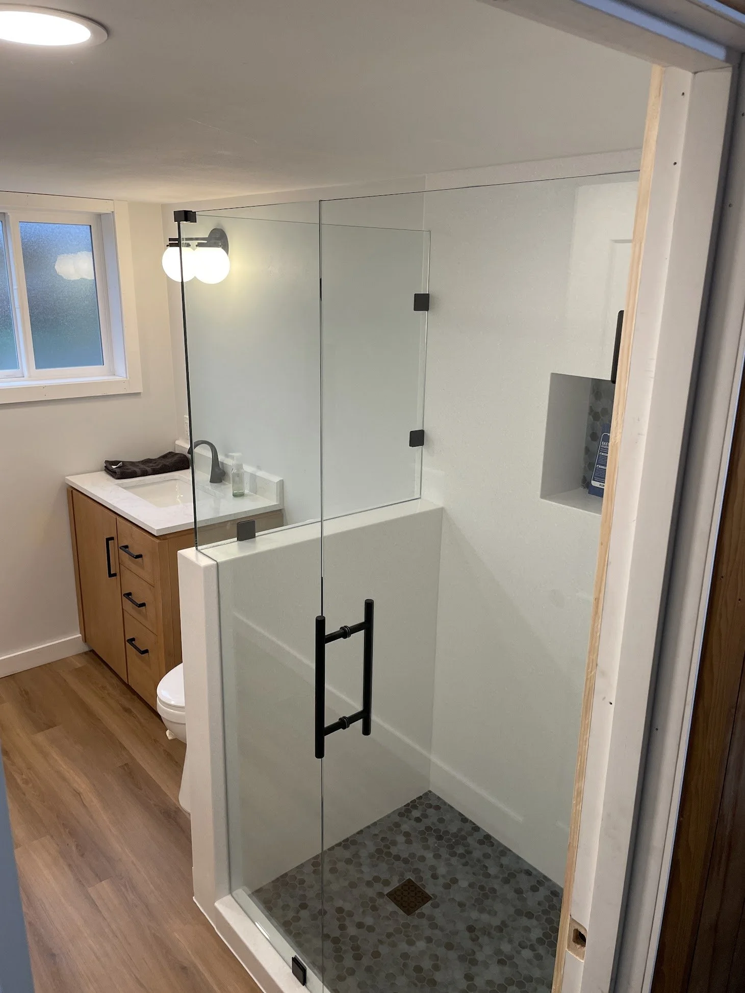 Bathroom with a walk-in shower that has clear glass walls, a small recessed shower niche, and patterned shower floor tiles. Adjacent is a vanity with a white countertop, a black faucet, and a towel. There is a window with frosted glass and a black wall sconce with two round bulbs.