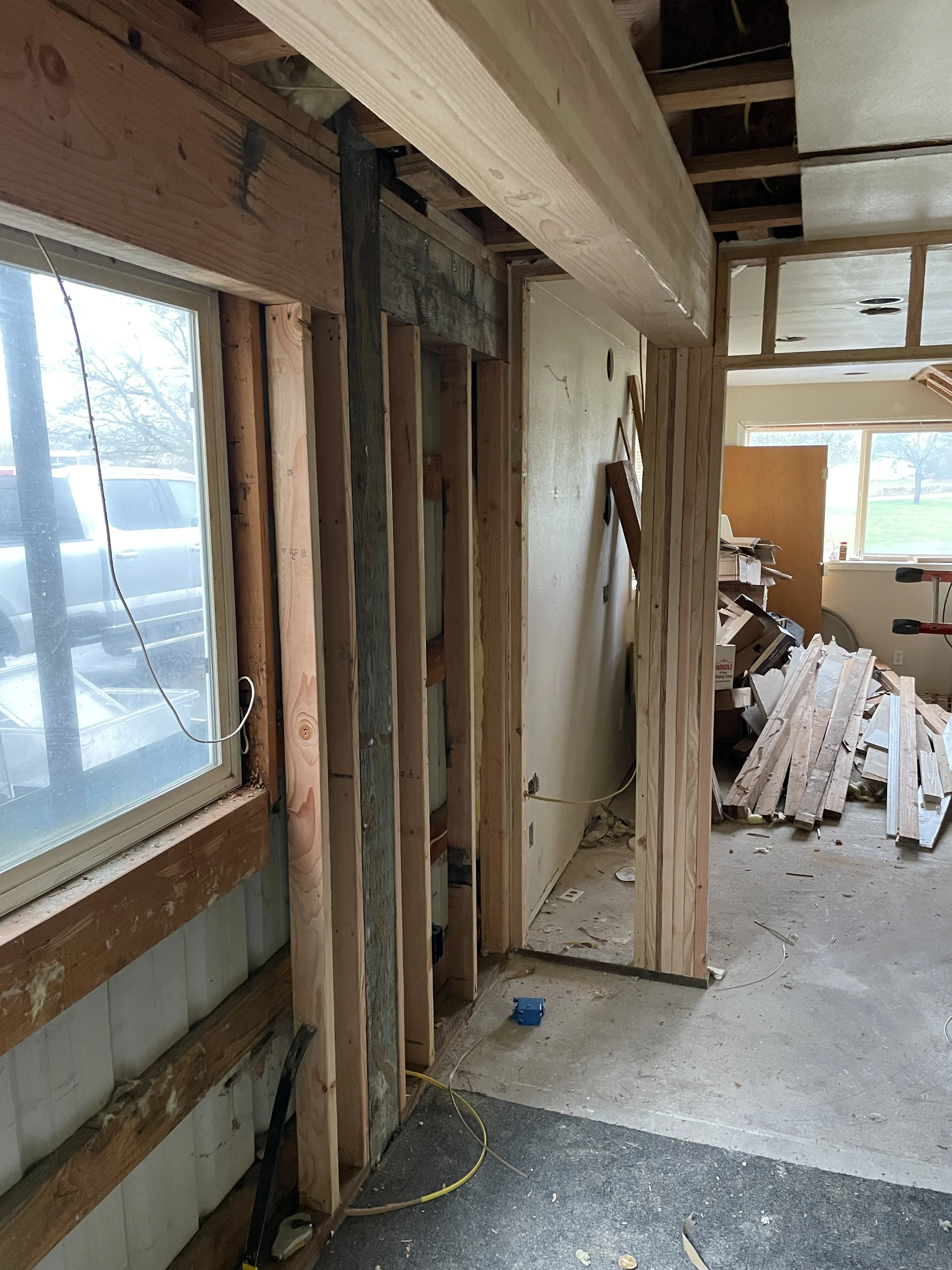 Interior view of a room under renovation with exposed wooden framing. The room has a window on the left side, and construction materials and debris on the floor. Piles of wooden planks and tools are visible in the background. Wooden beam.