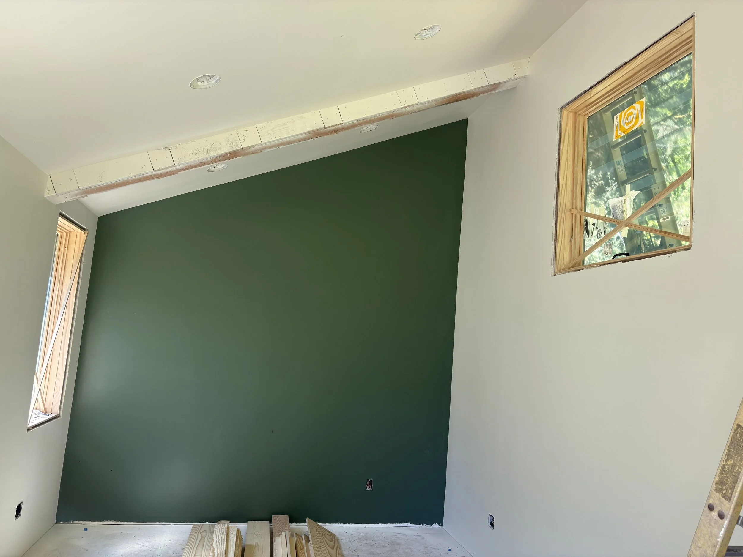 Interior of a room under construction with unfinished walls, a green accent wall, a window with wood trim, and electrical outlets and lights ceiling fixtures.
