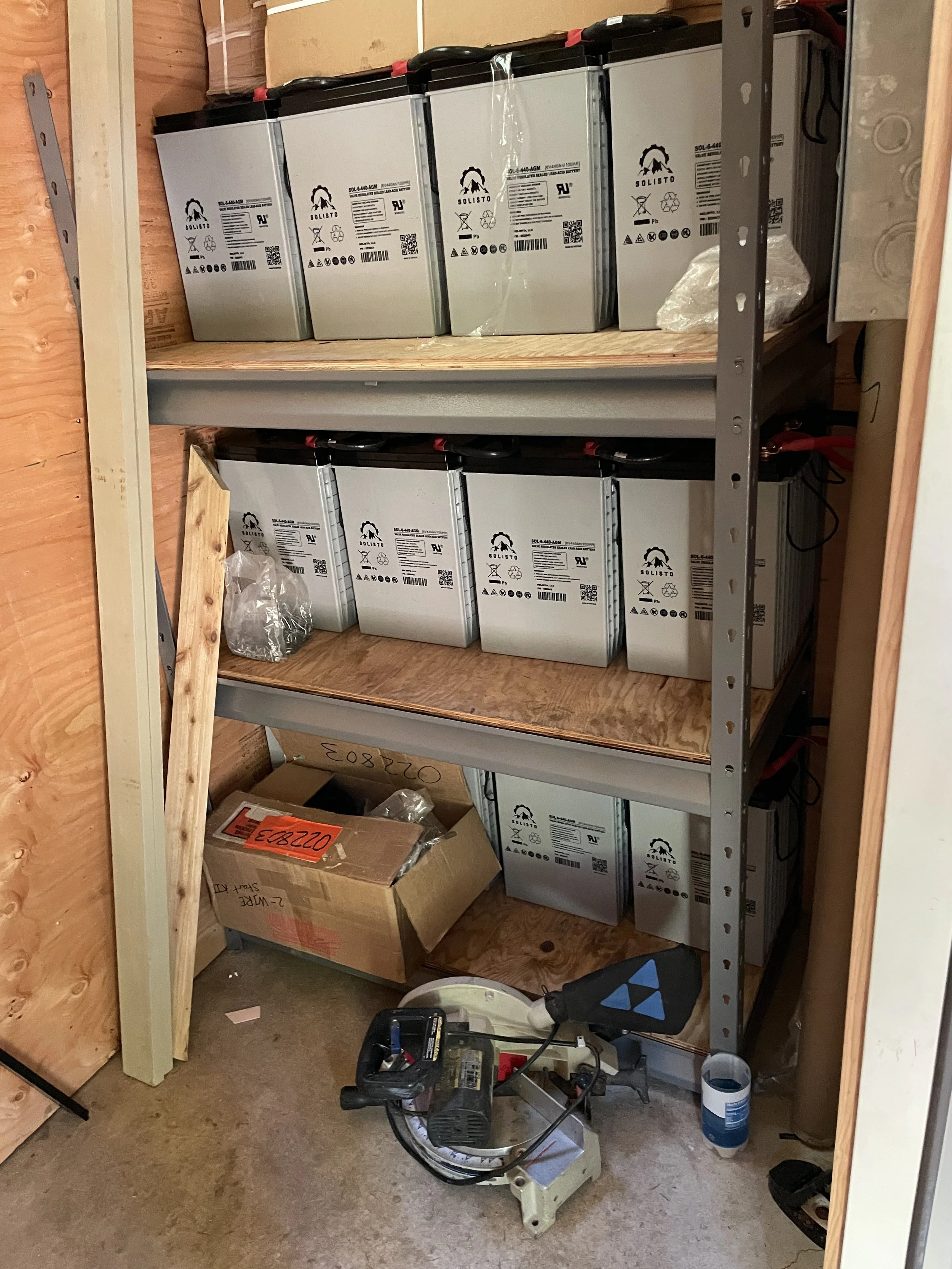 A storage room with metal shelves holding multiple batteries, a cardboard box, and various tools and equipment including a power saw and a red toolbox.