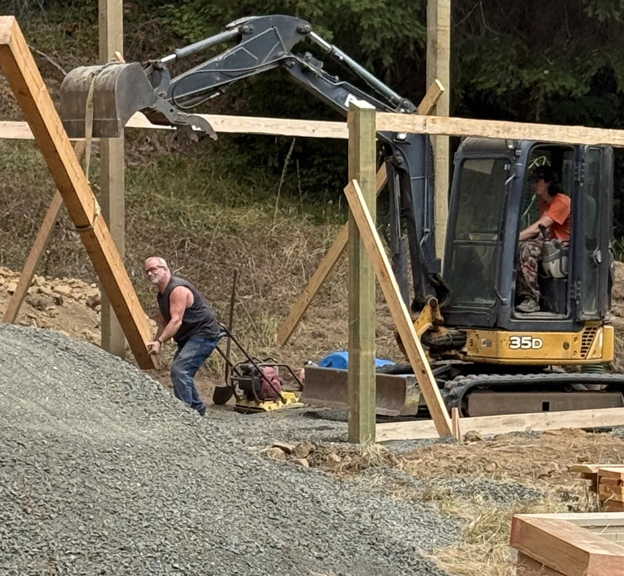 Construction site with a small yellow excavator and workers installing wooden fence posts.