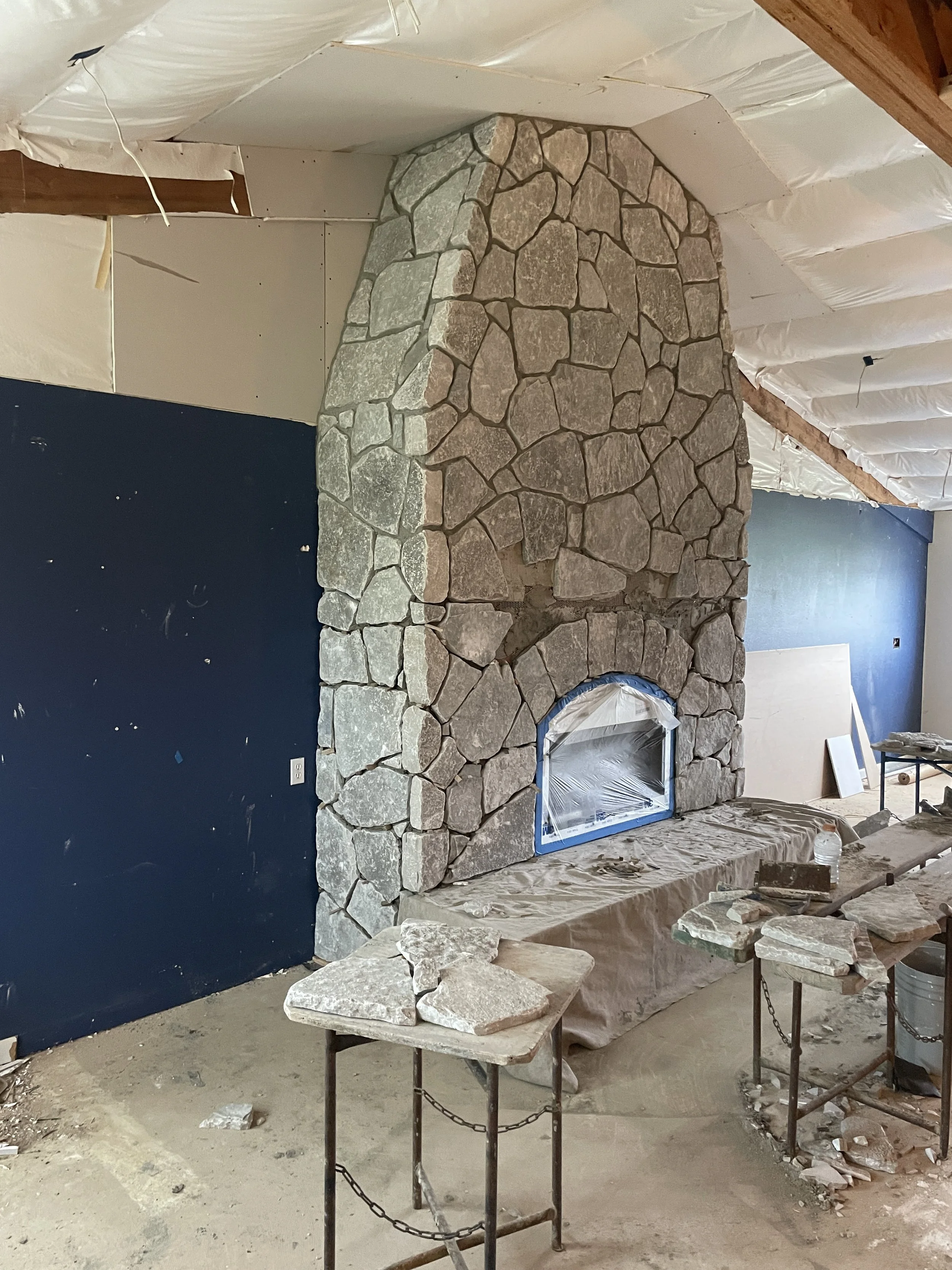 A stone fireplace under construction inside a room with partially finished white and blue walls, with construction tools and materials scattered around.