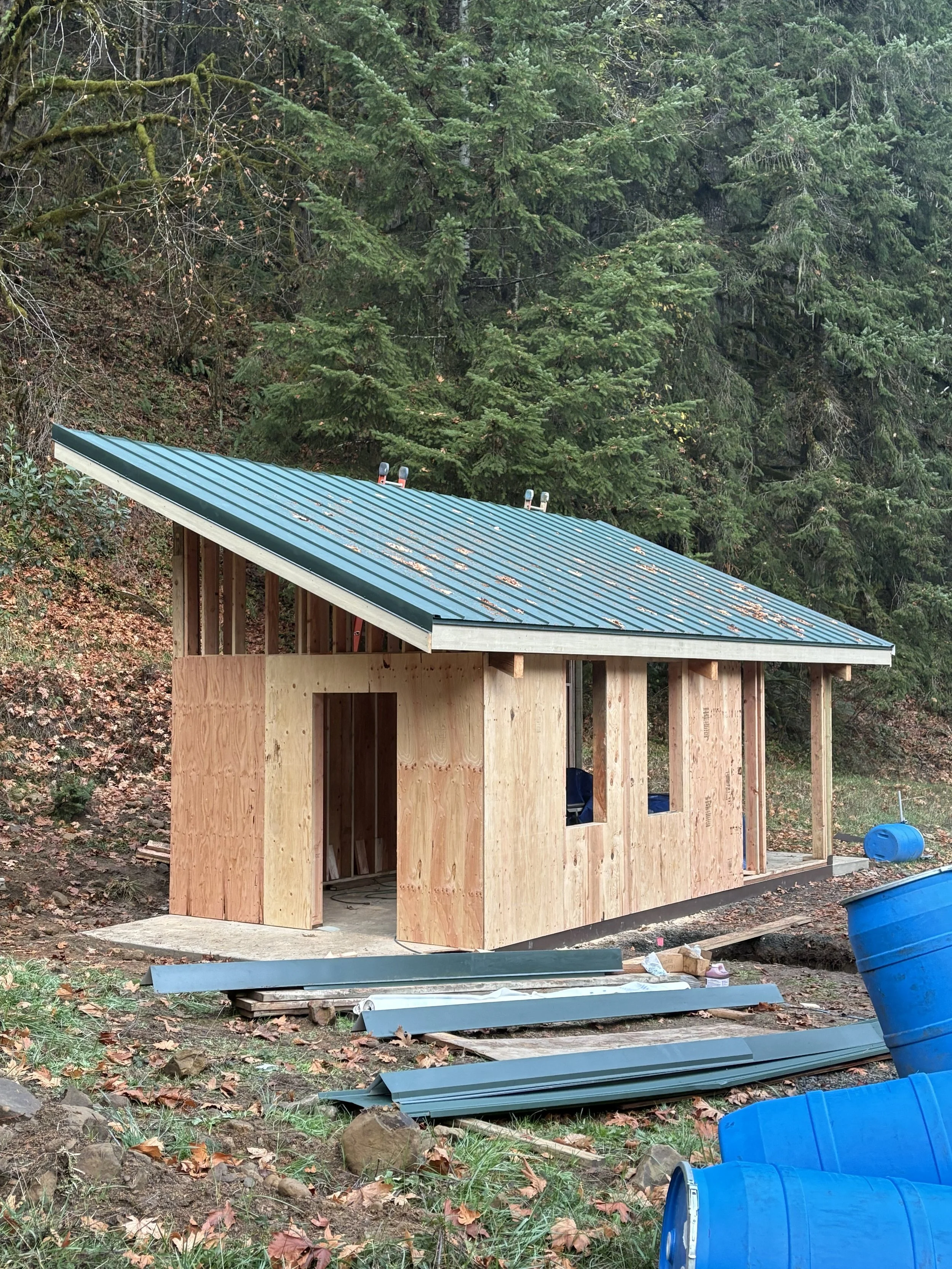 A small building under construction surrounded by a forested area, with a metal roof, wooden walls, and construction materials and barrels on the ground.