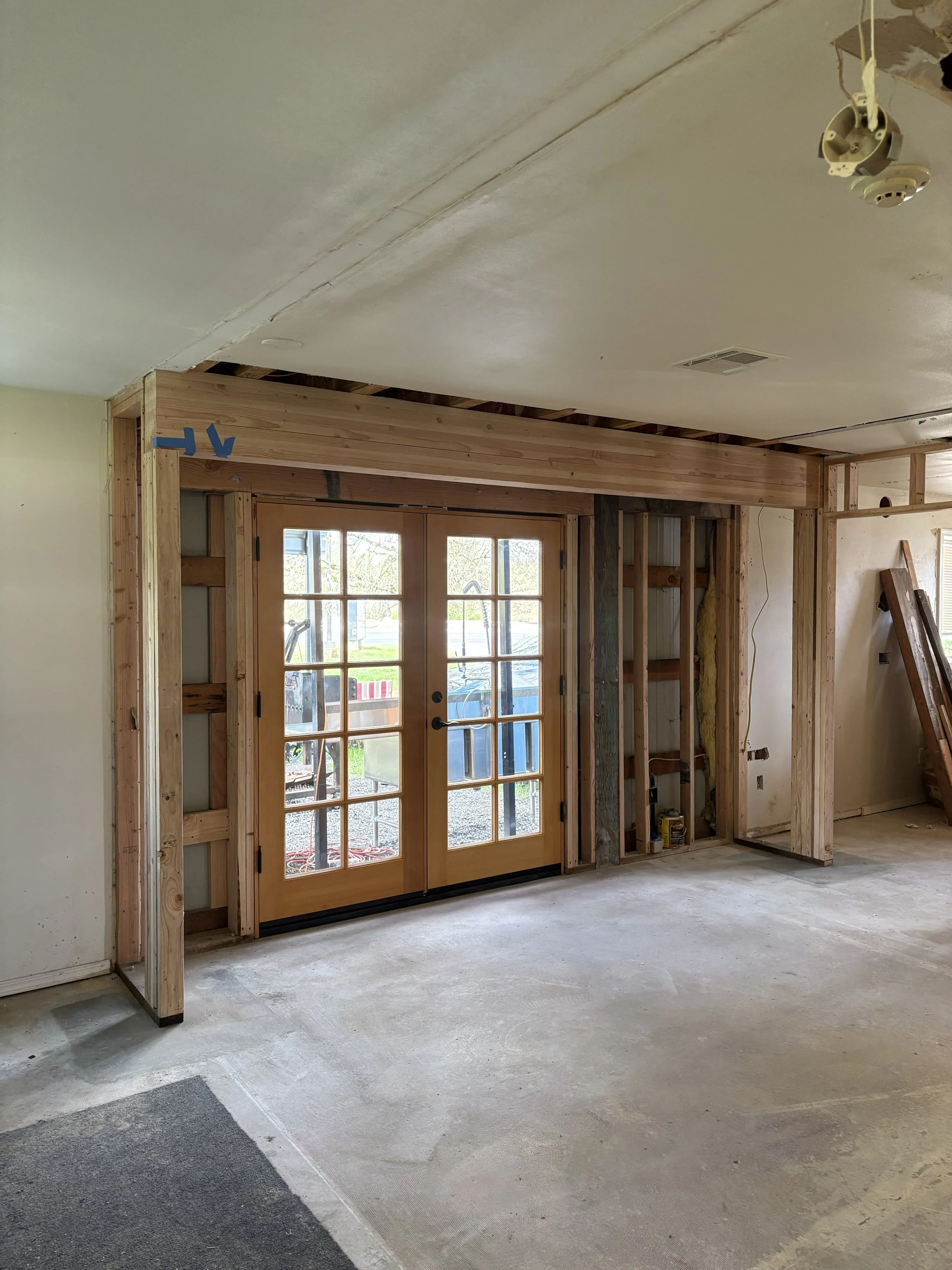 Construction site of a room with a double glass door, exposed wooden framing, and unfinished walls and ceiling.